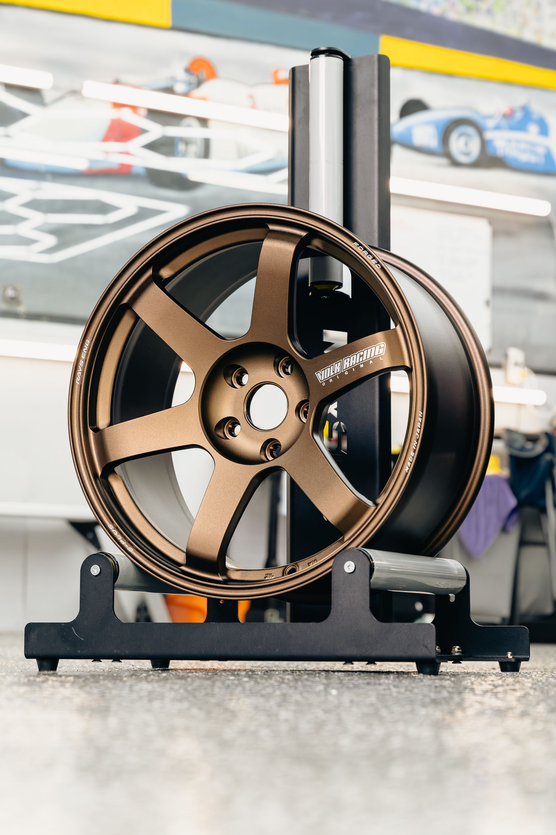 A bronze, six-spoke Rays Volk Racing alloy wheel sits mounted on a black balancing stand in a workshop.