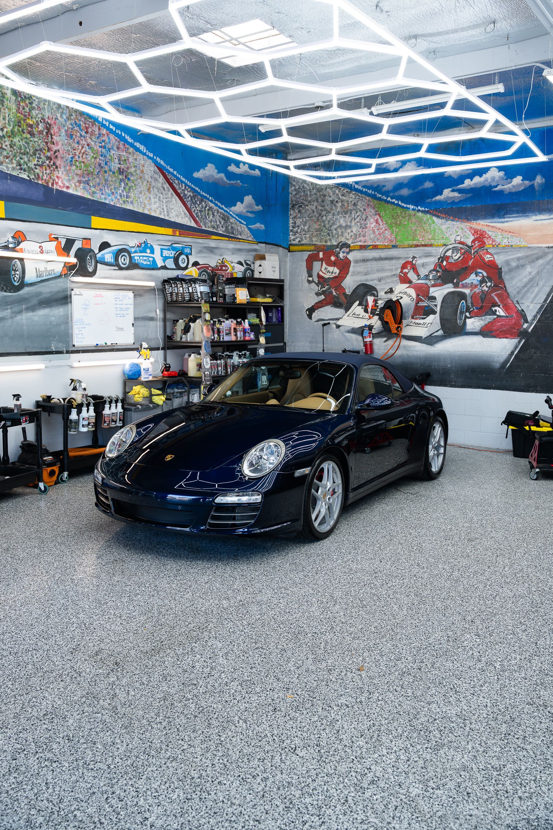A dark blue Porsche parked in a garage with a hexagonal LED ceiling light and a mural of race cars on the walls.