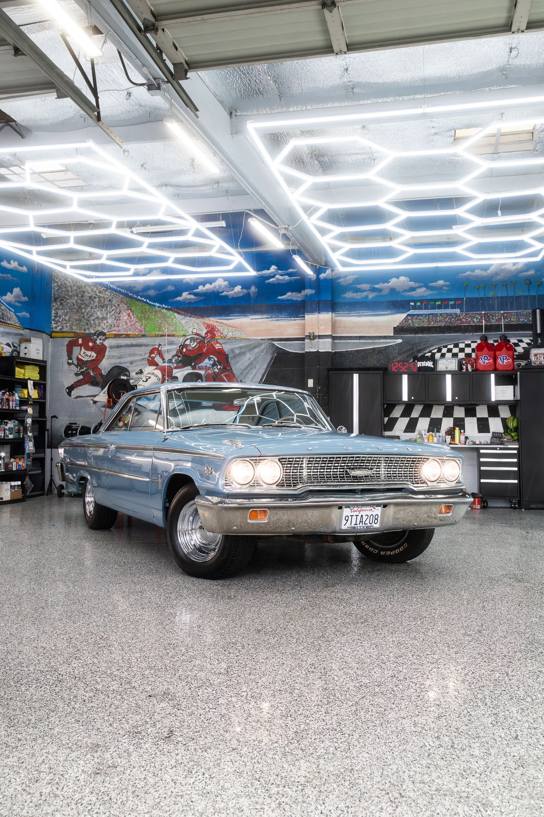 A classic light blue convertible parked inside a garage with patterned floor and illuminated hexagonal ceiling panels.