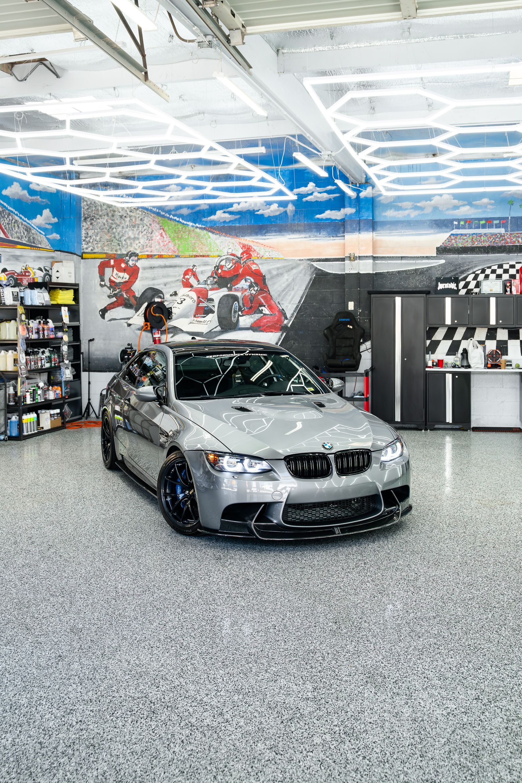 A silver BMW parked inside a garage featuring a mural of a race track and modern hexagonal ceiling lighting.