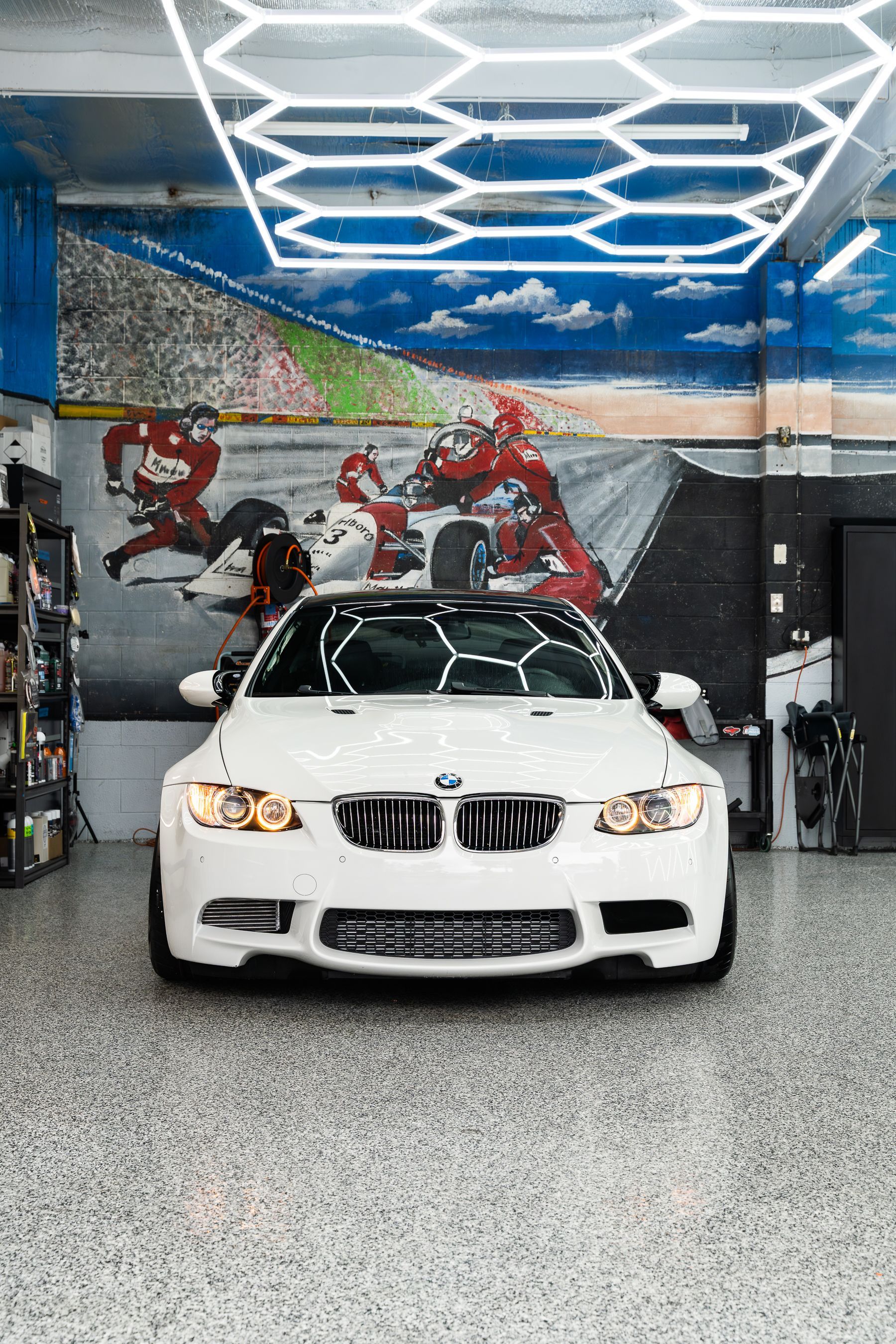 A white BMW sports car parked in a garage with a racing-themed mural and modern hexagonal ceiling lights.