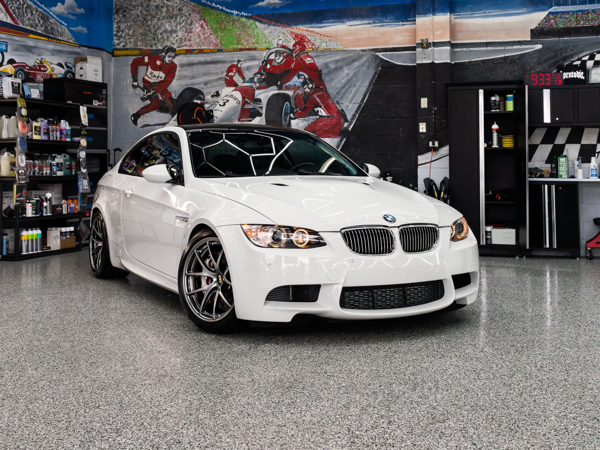 White BMW M3 coupe parked inside a well-lit garage with racing-themed wall art and storage shelves.