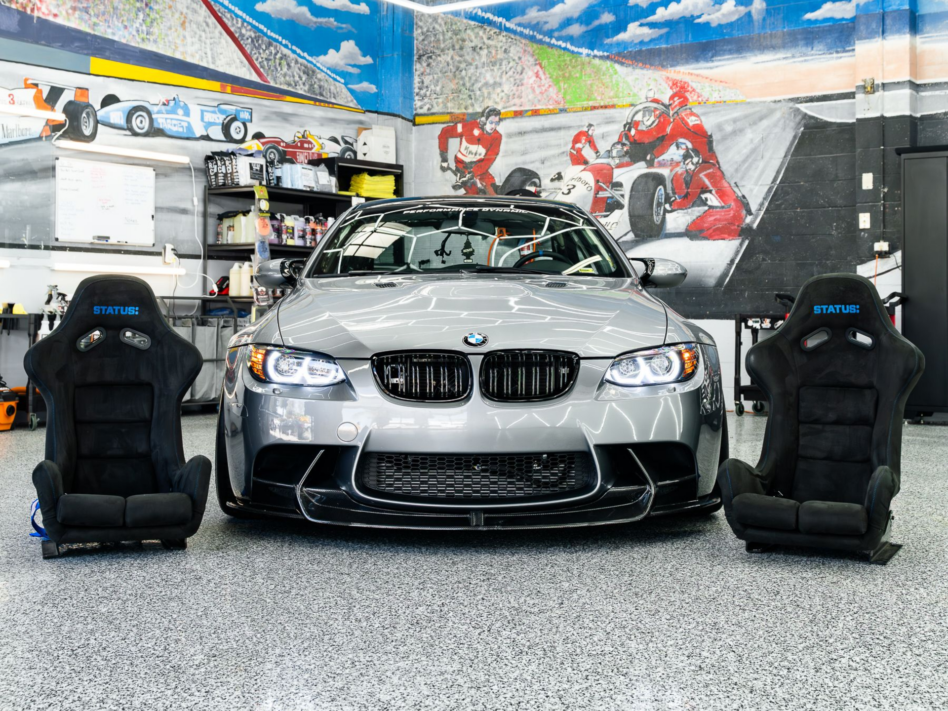 A silver BMW M3 parked in a garage, flanked by two black racing bucket seats on a speckled floor.