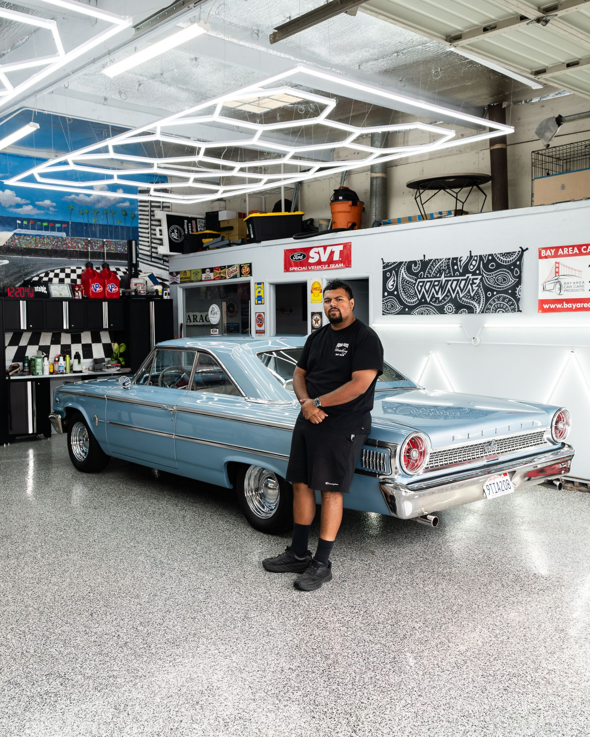 A person in a black t-shirt leans against a vintage light-blue car in a brightly lit, professional garage.