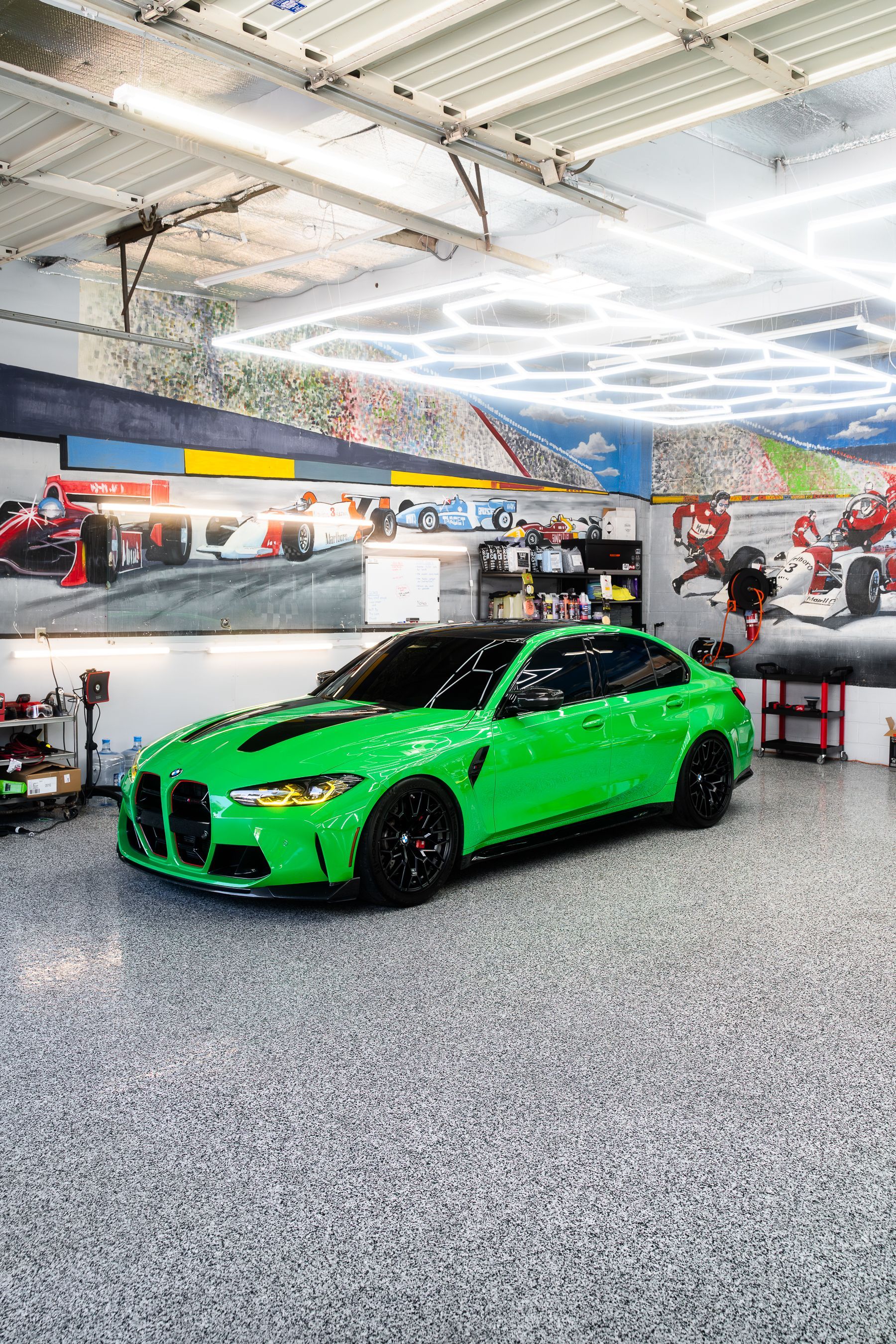 A white BMW sports car parked in a garage with a racing-themed mural and modern hexagonal ceiling lights.
