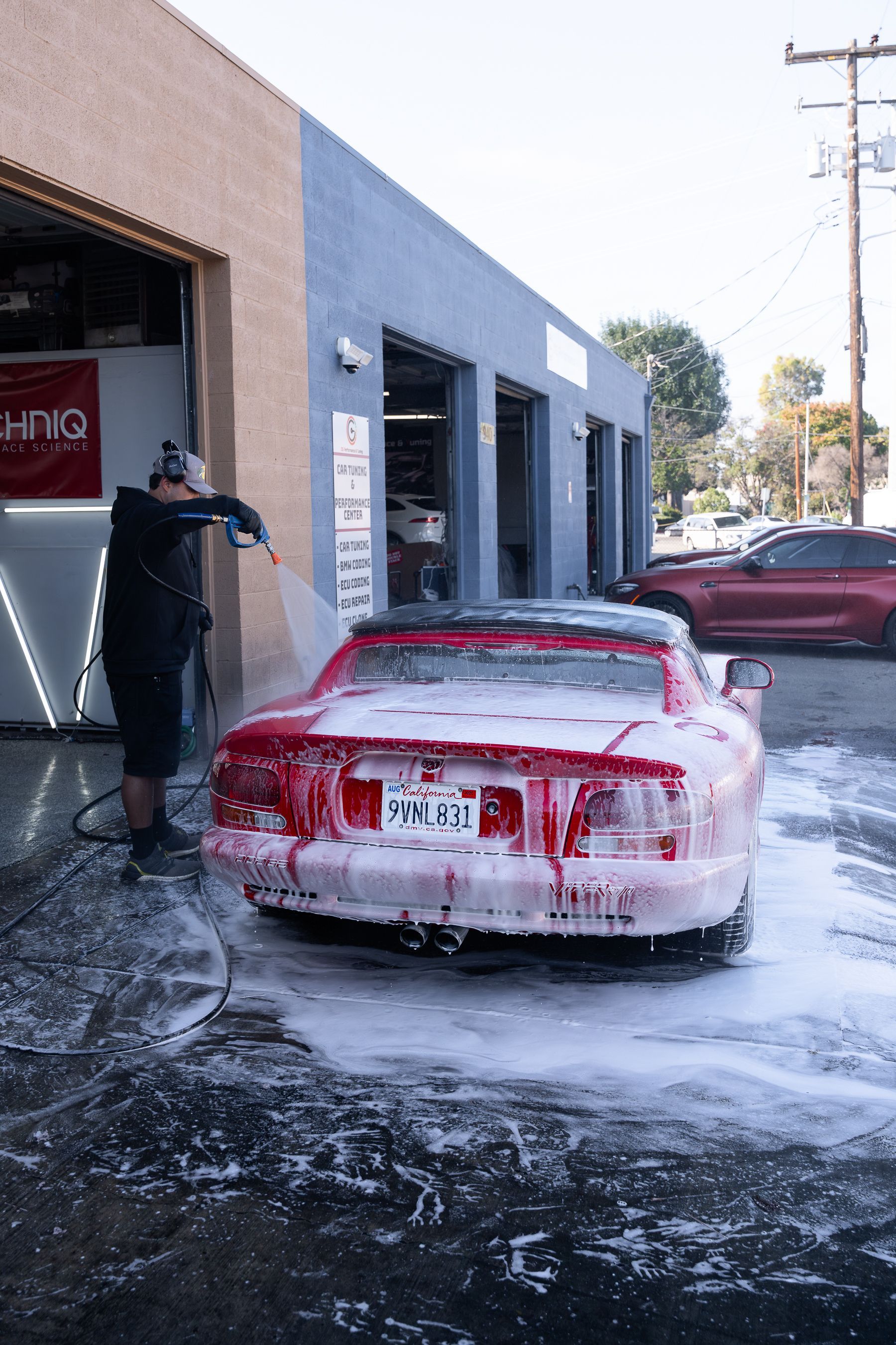 A technician pressure-washes a red sports car covered in soap suds outside a garage.