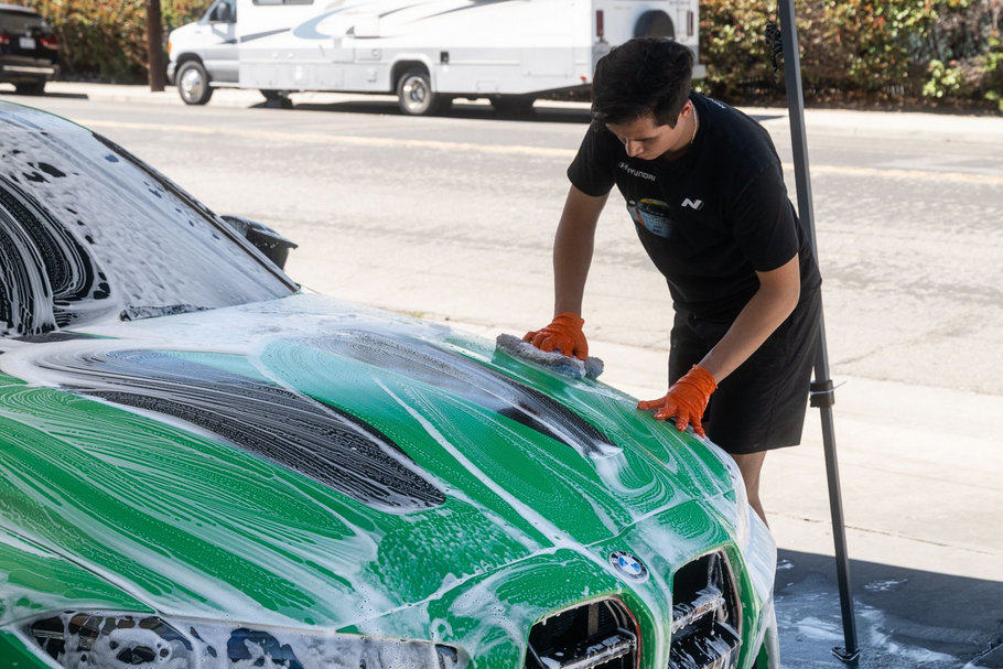 A person wearing orange gloves soaps and hand-washes the green hood of a car outdoors.