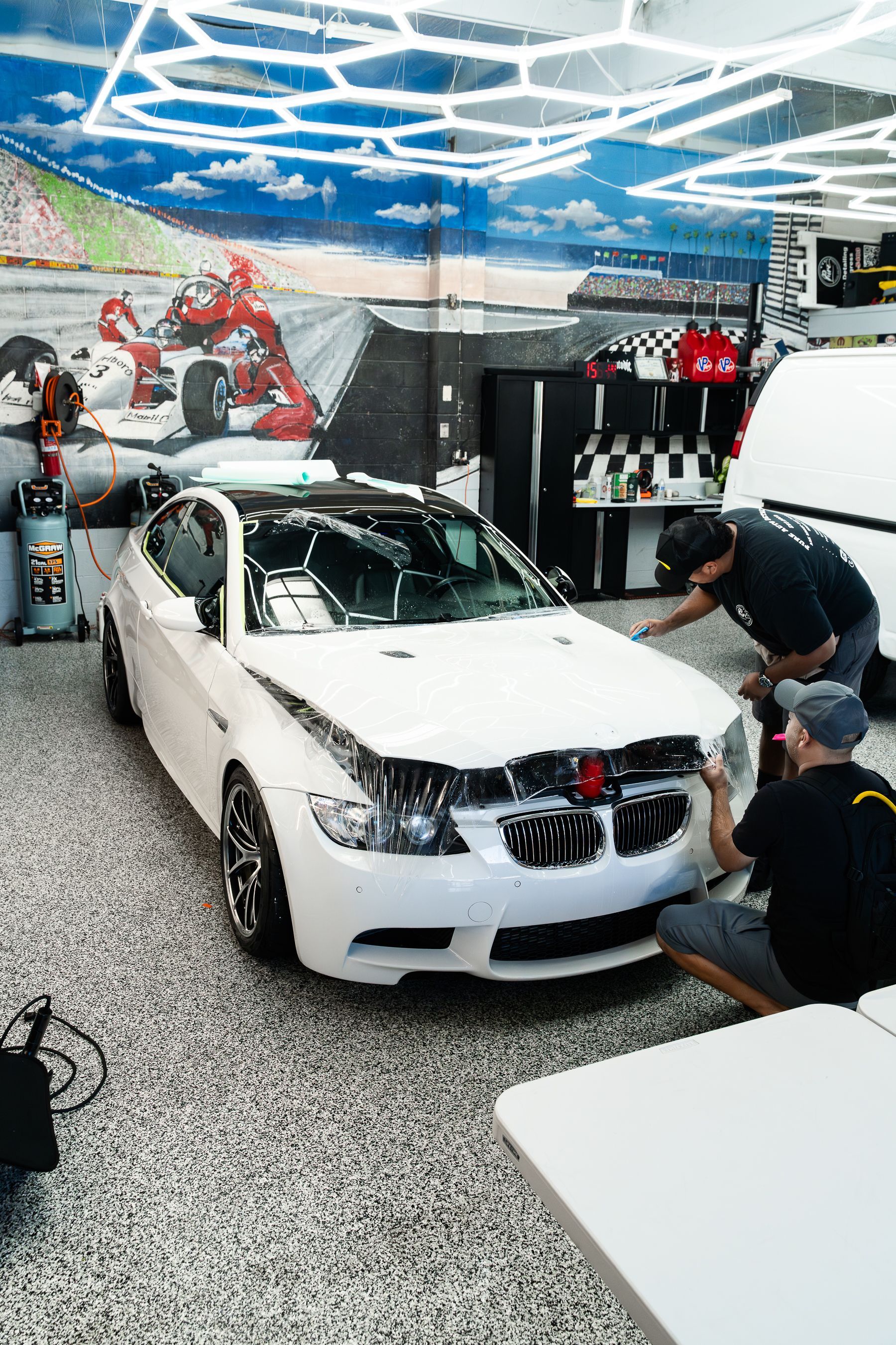 Two people work on applying a black protective film to the front bumper of a white BMW sedan in a garage.