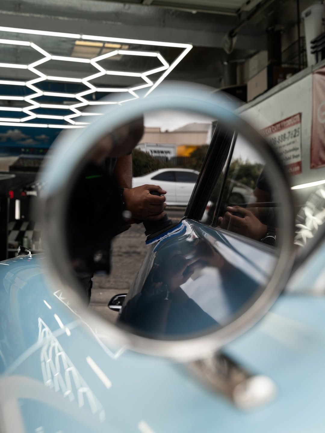 A close-up view through a round car side mirror showing a person working on a light blue car in a garage with hexagon lights.