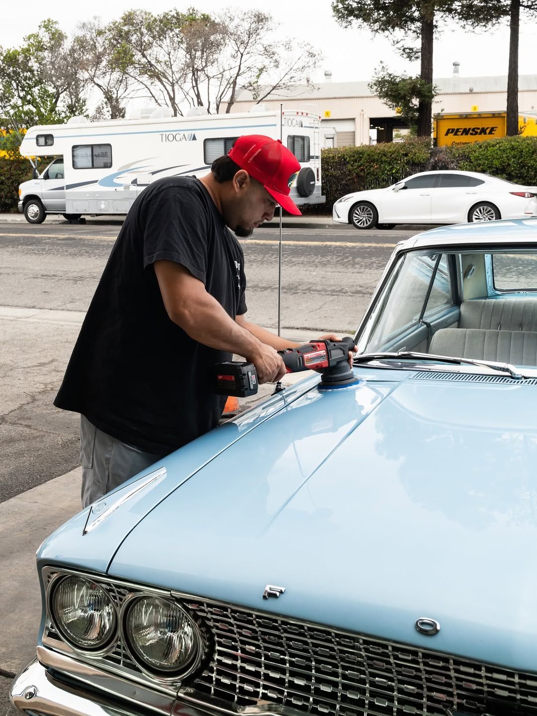 A person in a red cap uses a power polisher on the hood of a light blue vintage car in an outdoor parking area.