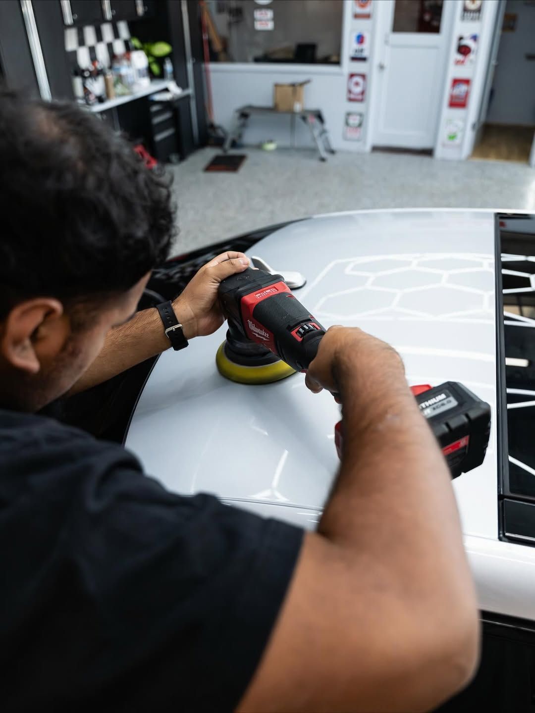 A person using a cordless power polisher on the white hood of a car inside a professional detailing shop.