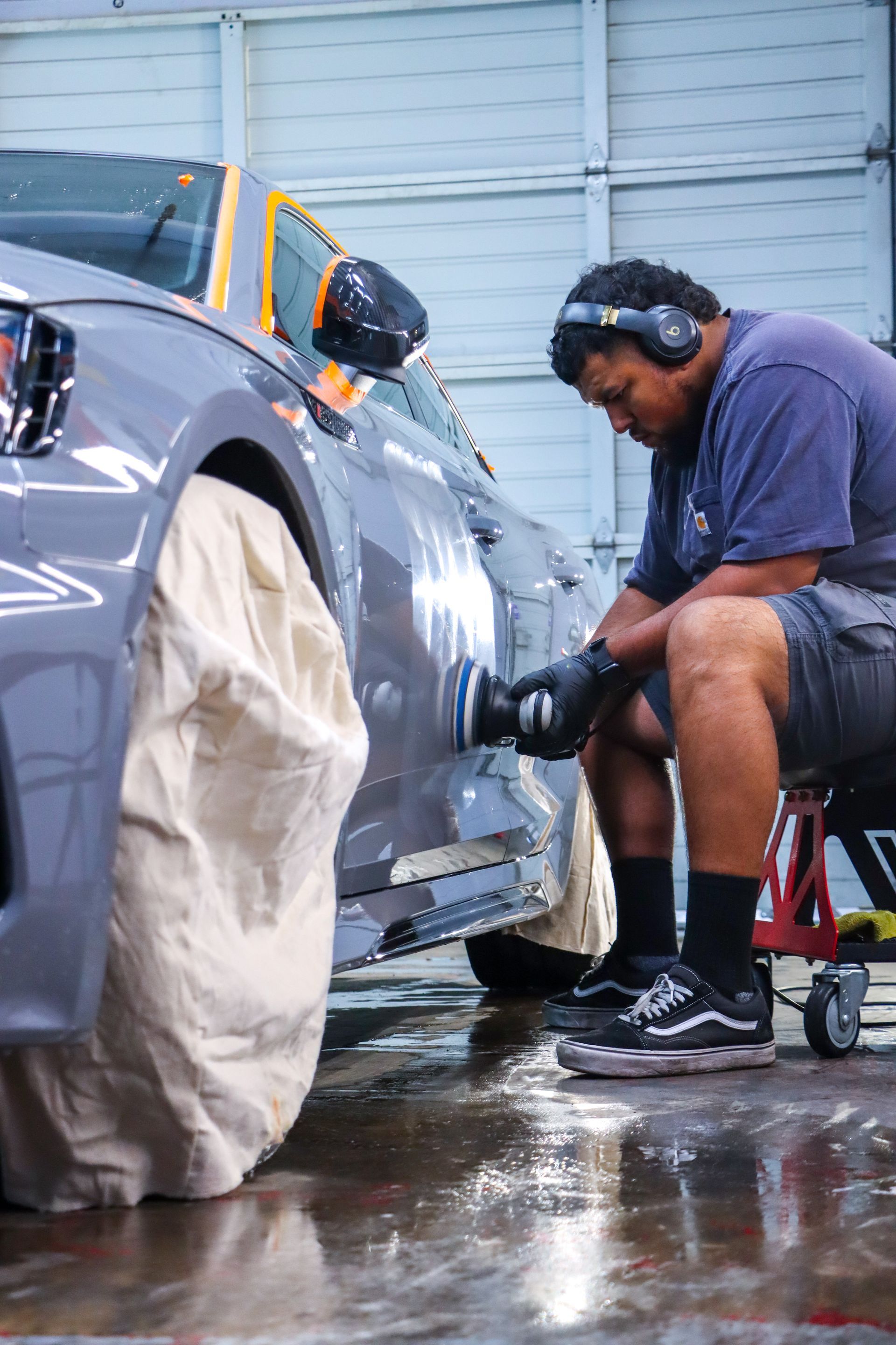 A technician in a workshop uses a power polisher on the grey side panel of a car covered with protective masking.
