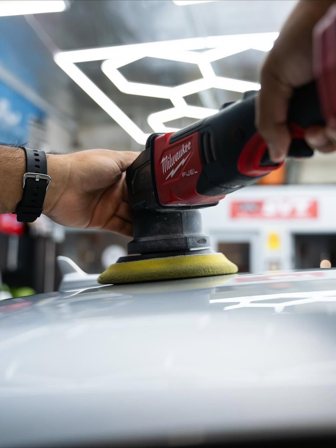 Hands use a red Milwaukee orbital polisher with a yellow pad to buff a car roof in a well-lit workshop.