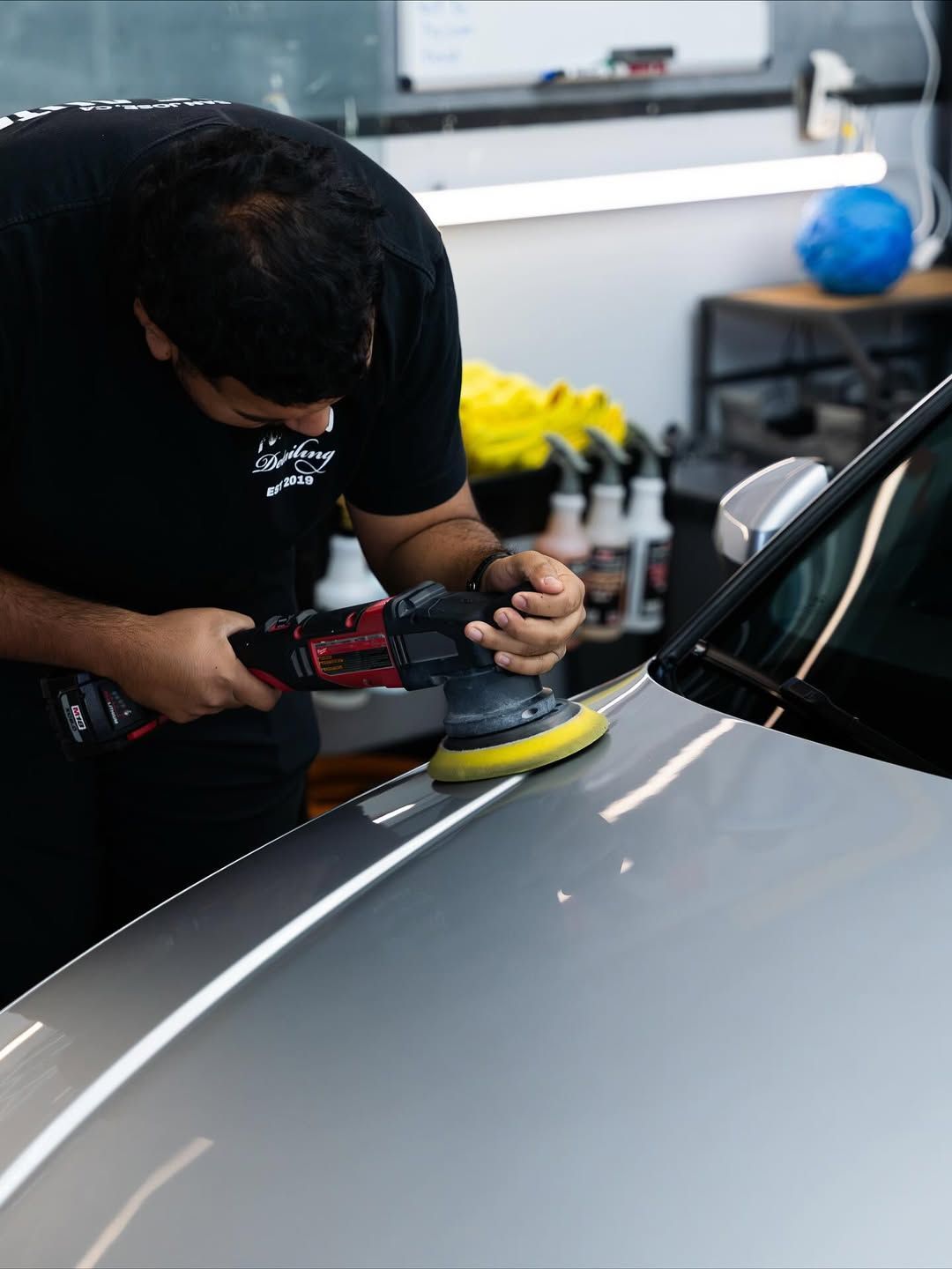 A person in a black t-shirt uses a power polisher on the grey hood of a car inside a brightly lit workshop.