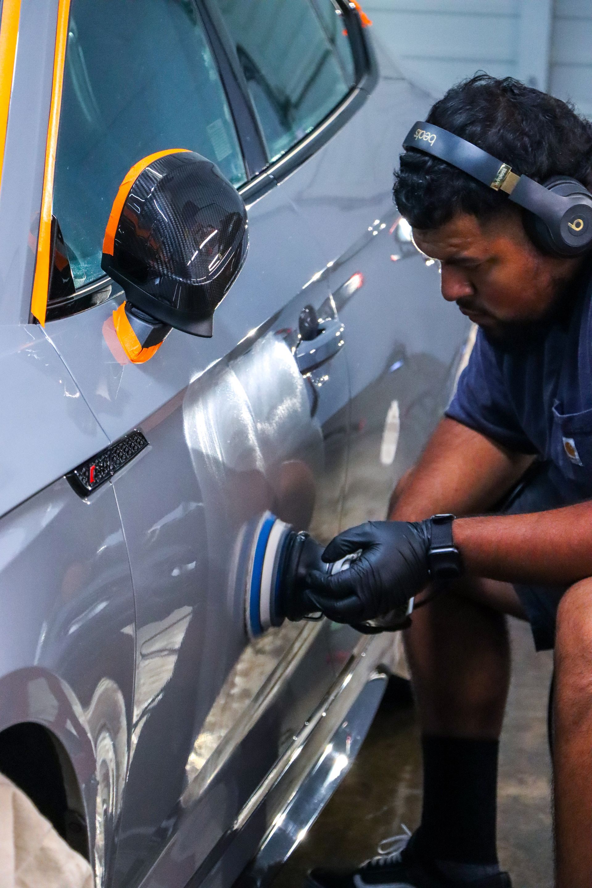 A professional detailer with headphones uses a power buffer to polish the grey door of a car in a workshop.