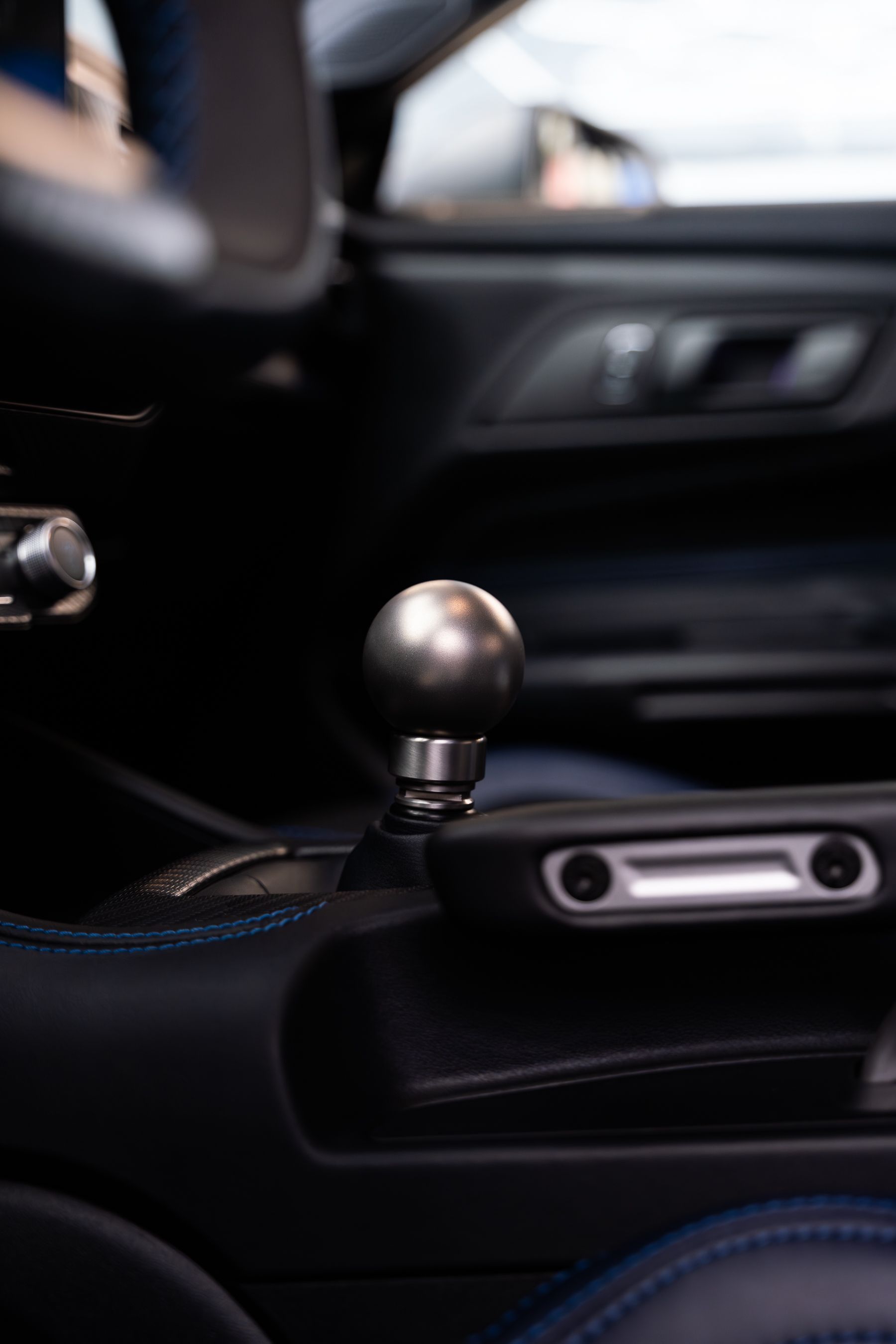 Close-up of a brushed metal spherical gear shifter in a dark car interior with blue stitching.