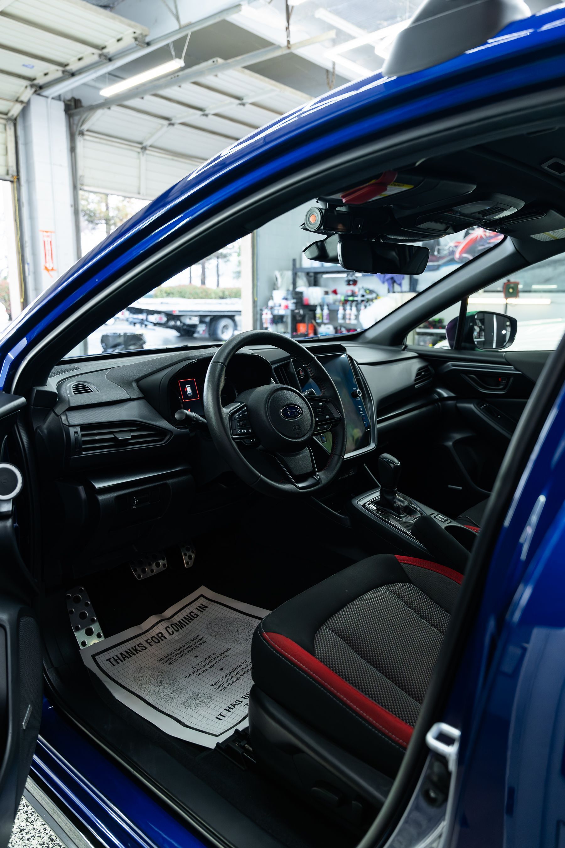 Interior of a blue car, showing the driver's seat with red trim, a black steering wheel, and a paper floor mat.