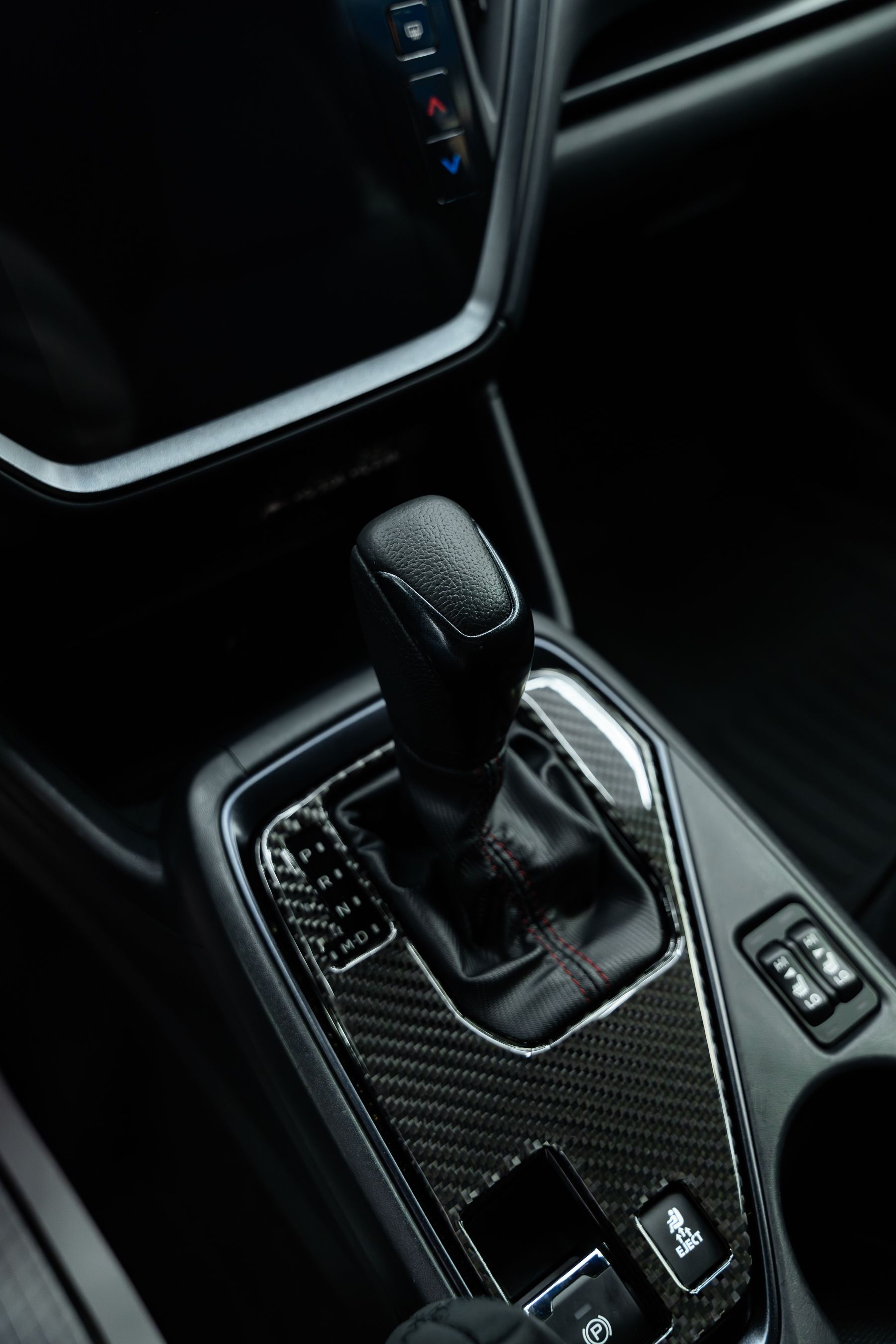 Interior of a car showing a black leather gear shifter surrounded by carbon fiber trim on the center console.