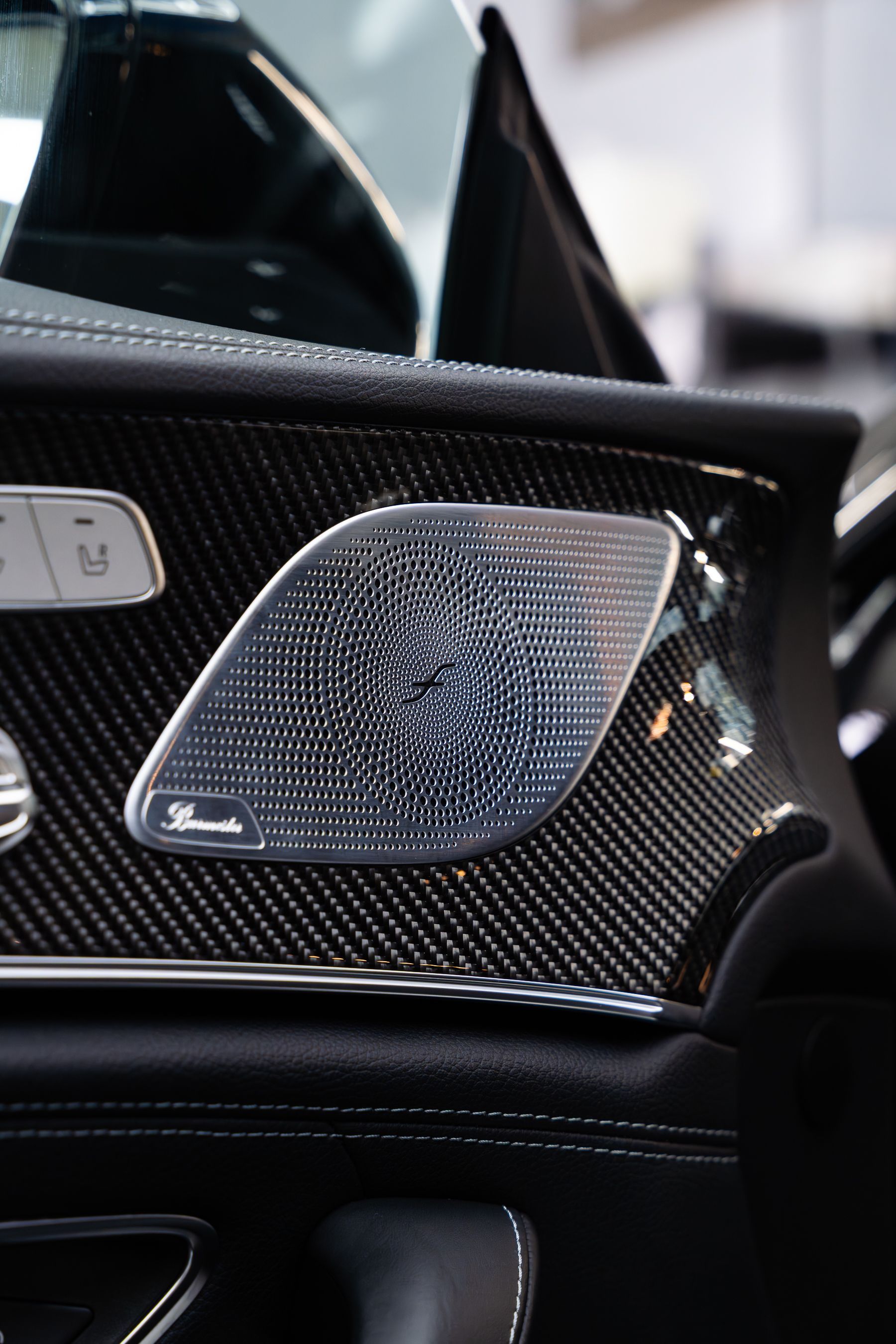 A close-up of a premium Burmester speaker grille set into a dark carbon fiber car door panel.