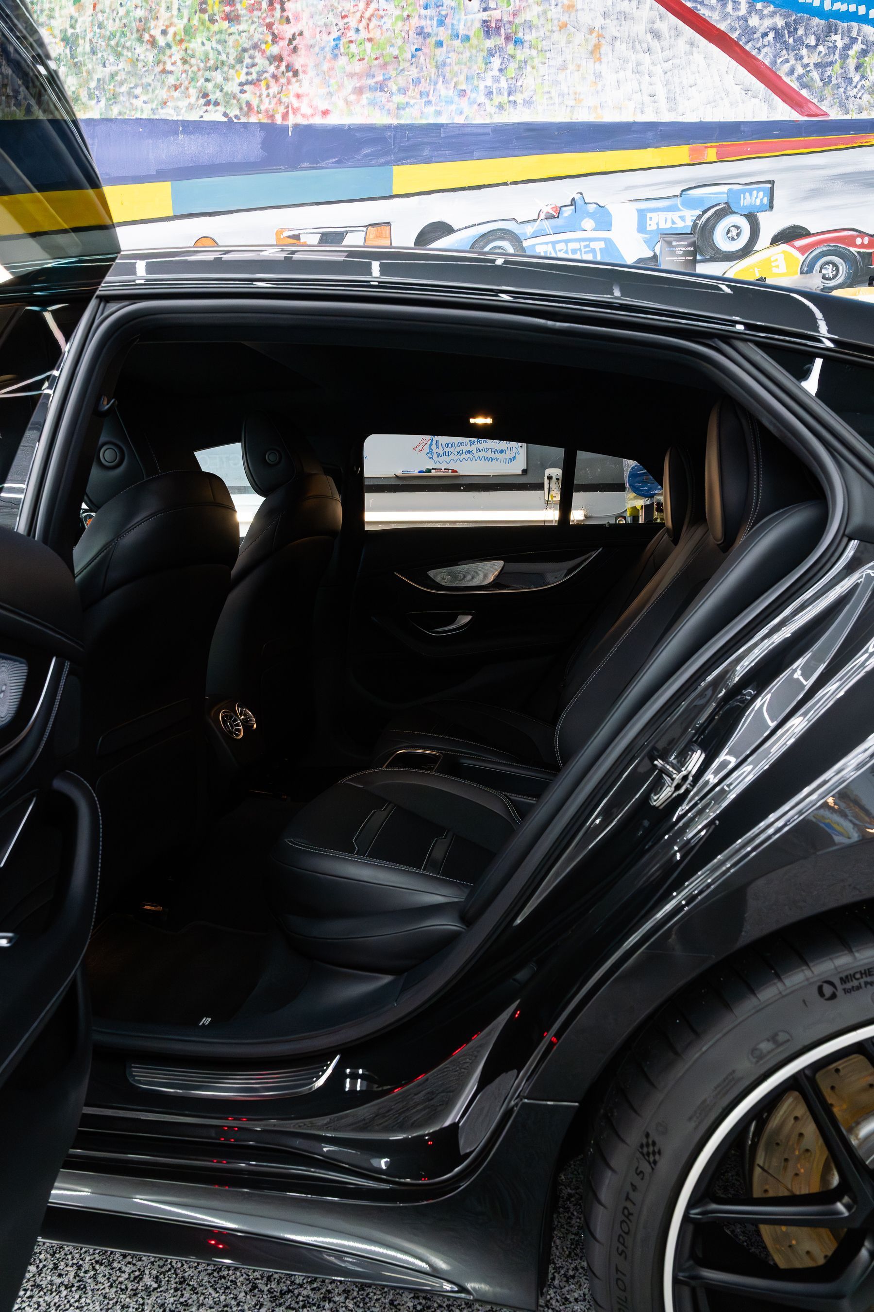 Open rear door of a black luxury car with perforated leather seats, parked in front of a colorful racing mural.