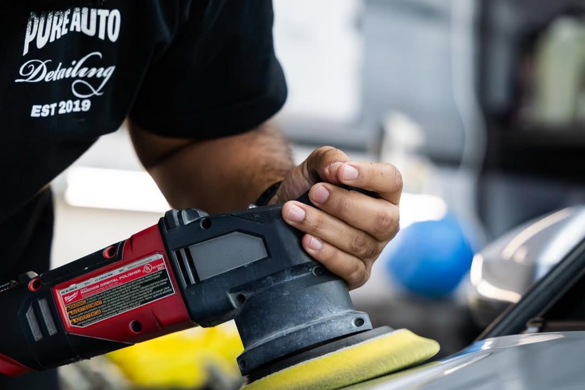 A person uses a red and black power buffer with a yellow pad to polish a vehicle's surface in a shop setting.