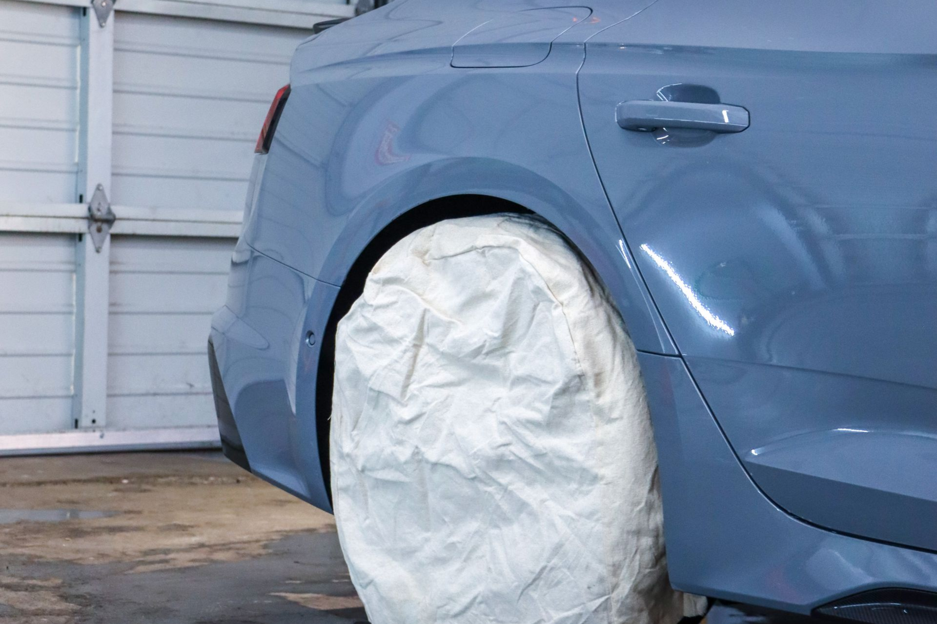 A blue car parked in a garage with its rear tire covered in a white protective bag for painting or maintenance.
