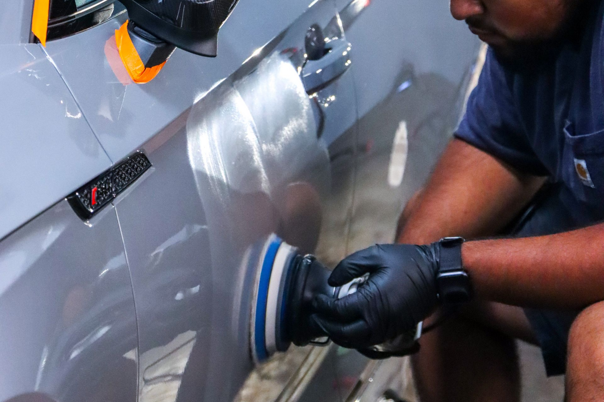A technician wearing black gloves uses a power polisher on the grey side panel of a vehicle.