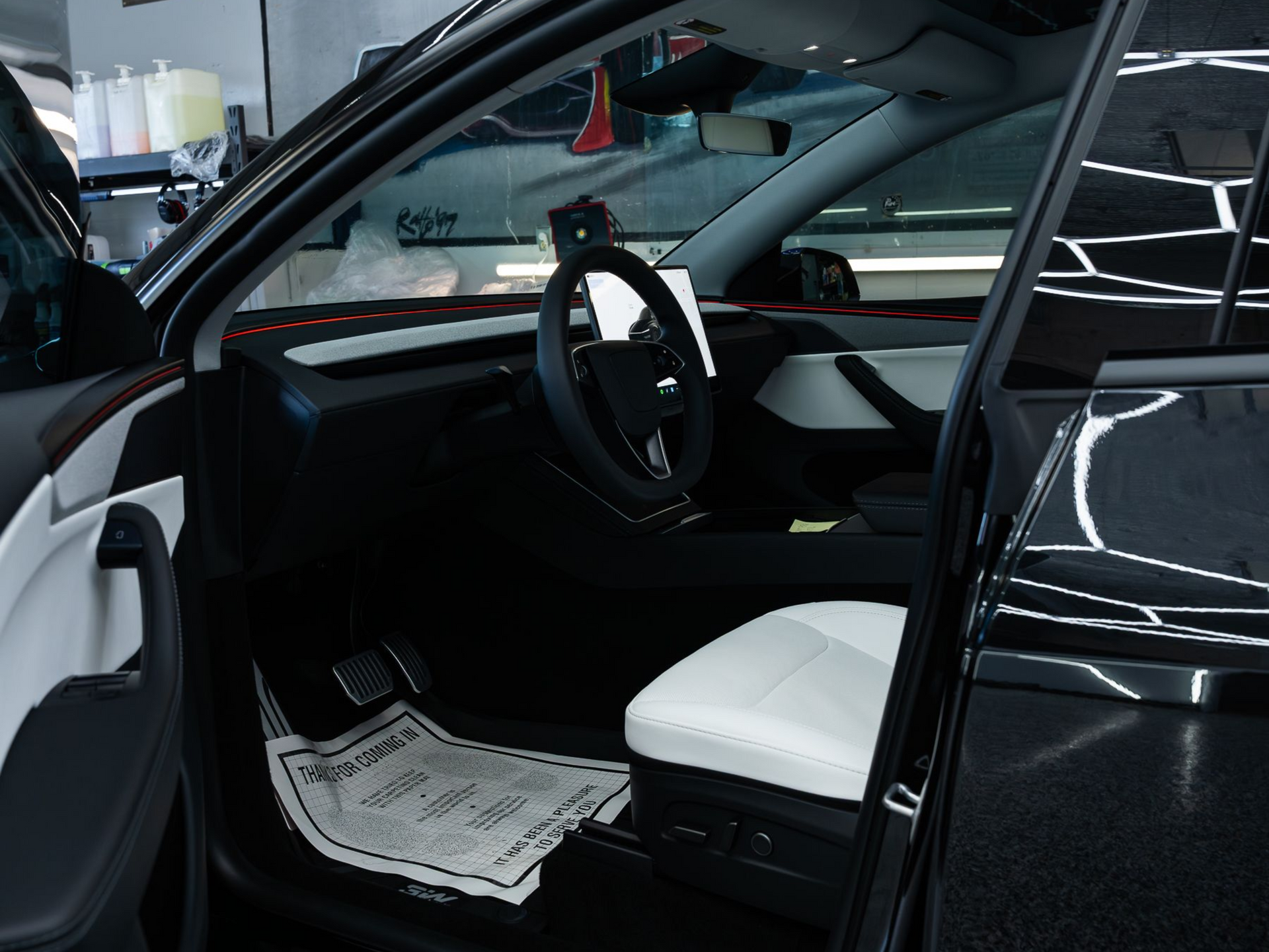 The interior of a black car with white leather seats, a steering wheel, and a protective floor mat.