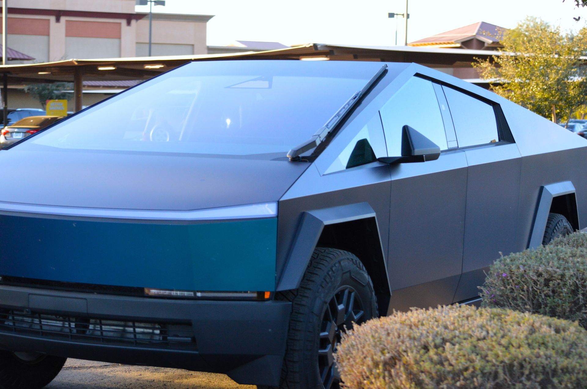A matte black Tesla Cybertruck with a triangular silhouette parked outdoors next to small bushes.