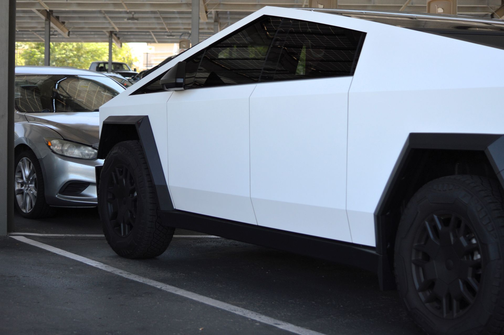 A white Tesla Cybertruck parked next to a silver sedan under a covered parking structure.