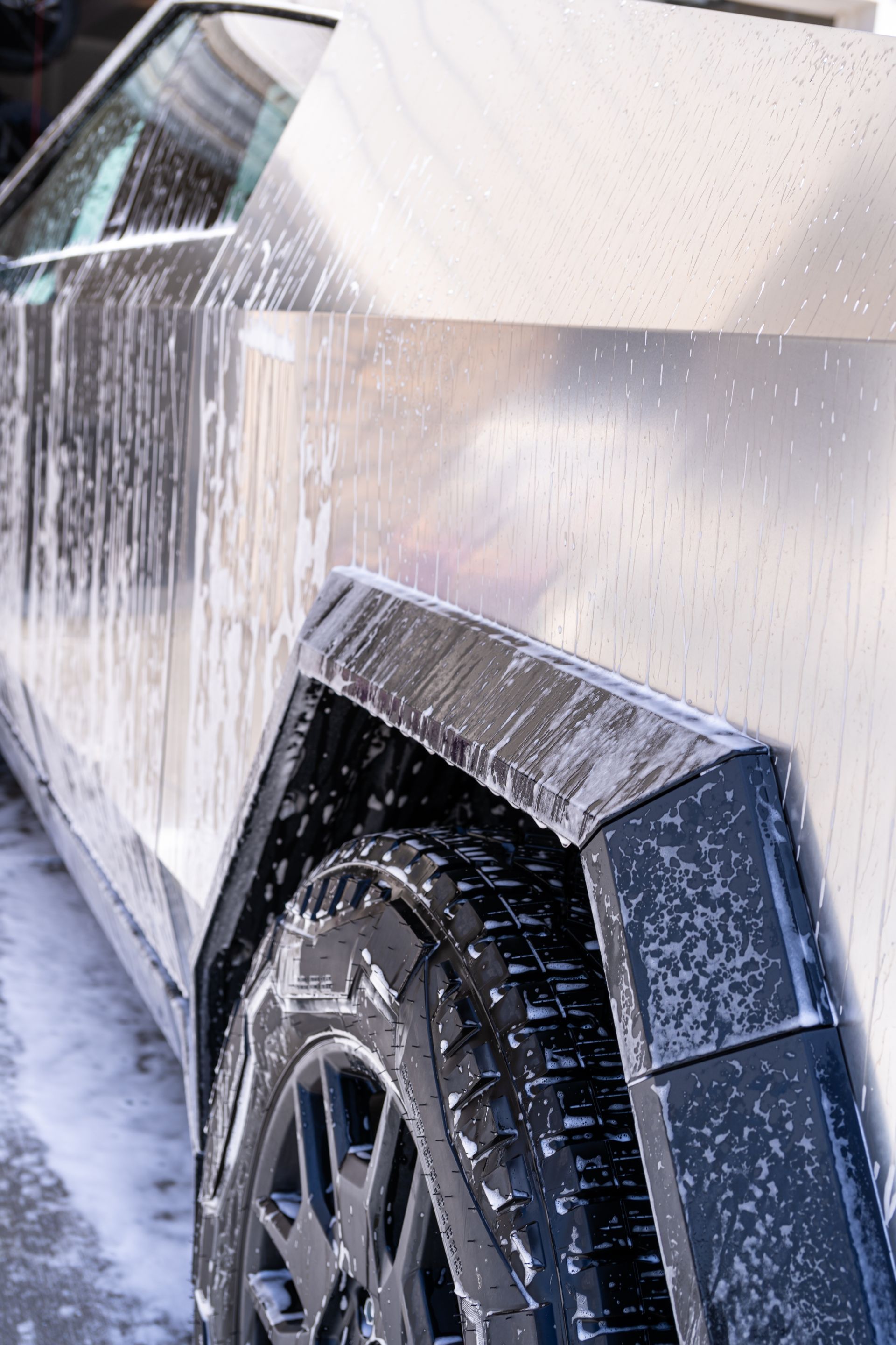 A close-up of a stainless steel Tesla Cybertruck covered in white soap suds during a car wash.