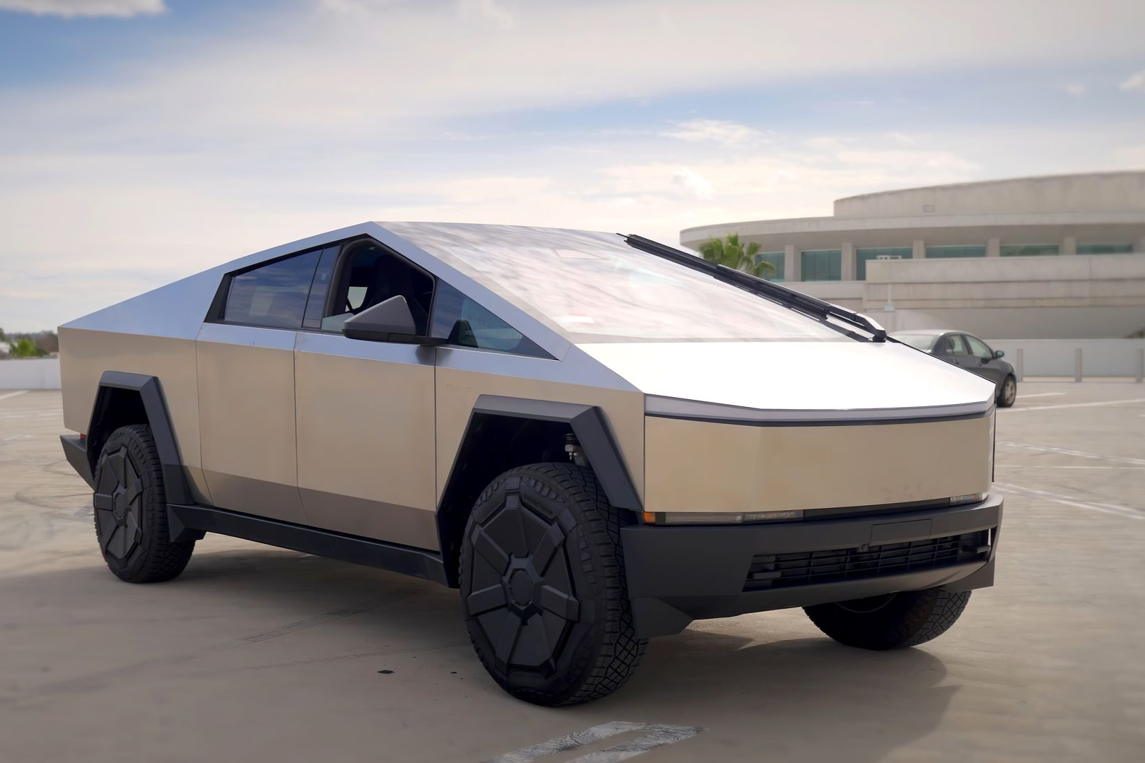A silver, angular Tesla Cybertruck parked on a concrete lot under a bright, cloudy sky.
