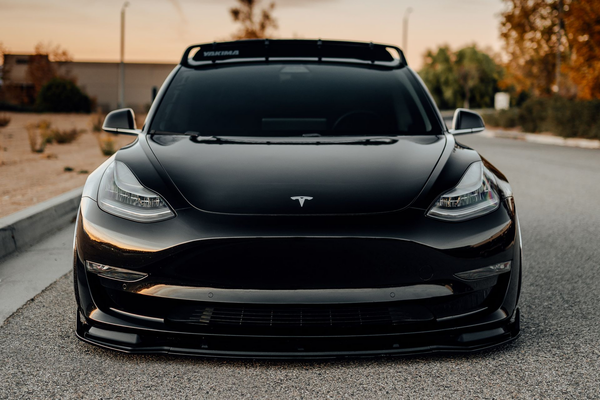 A modified black Tesla Model 3 with a front lip spoiler and roof rack, parked on a paved road during sunset.