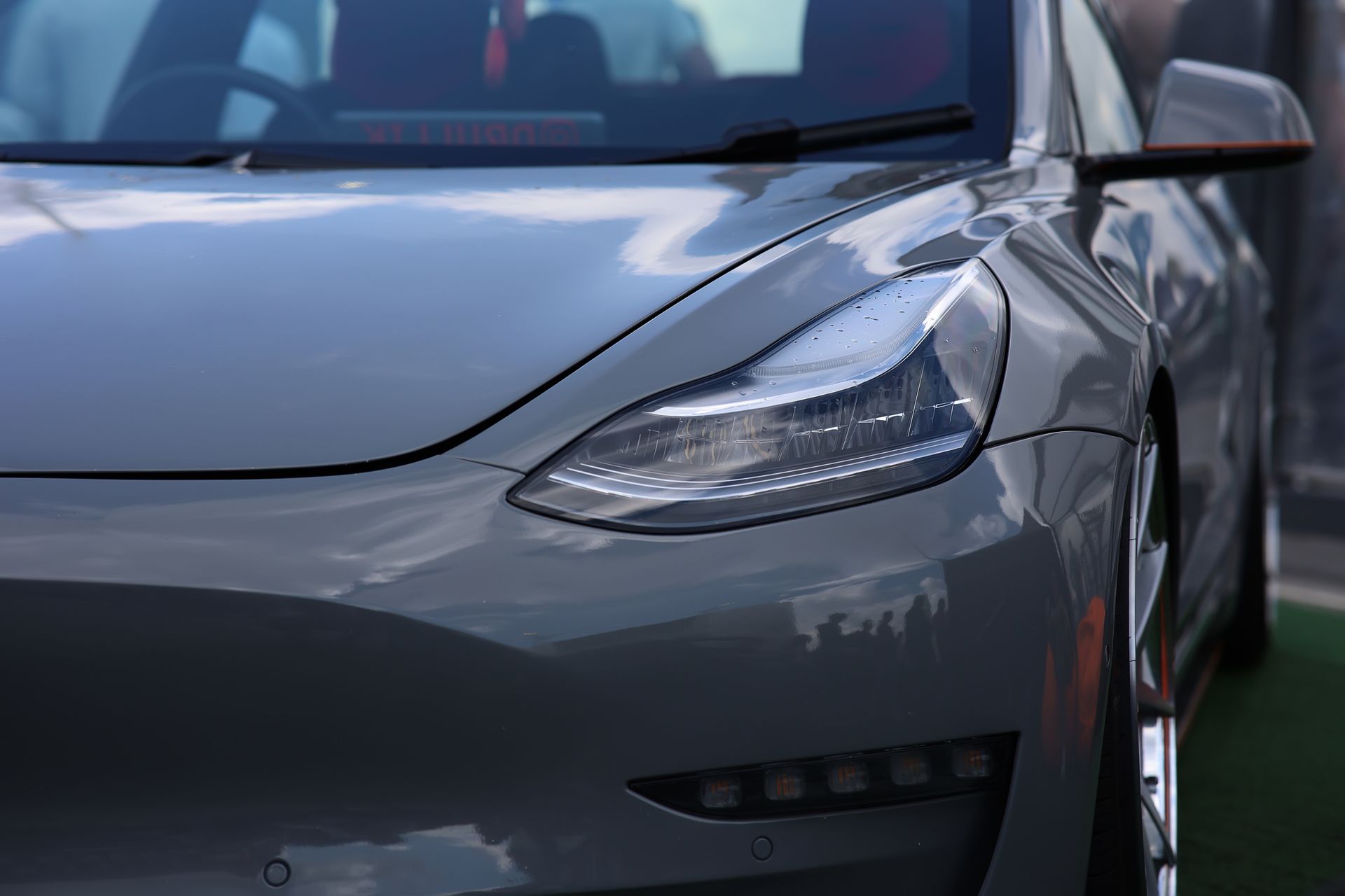Close-up of a grey Tesla Model 3 front end, highlighting the headlight assembly and fender on a bright day.