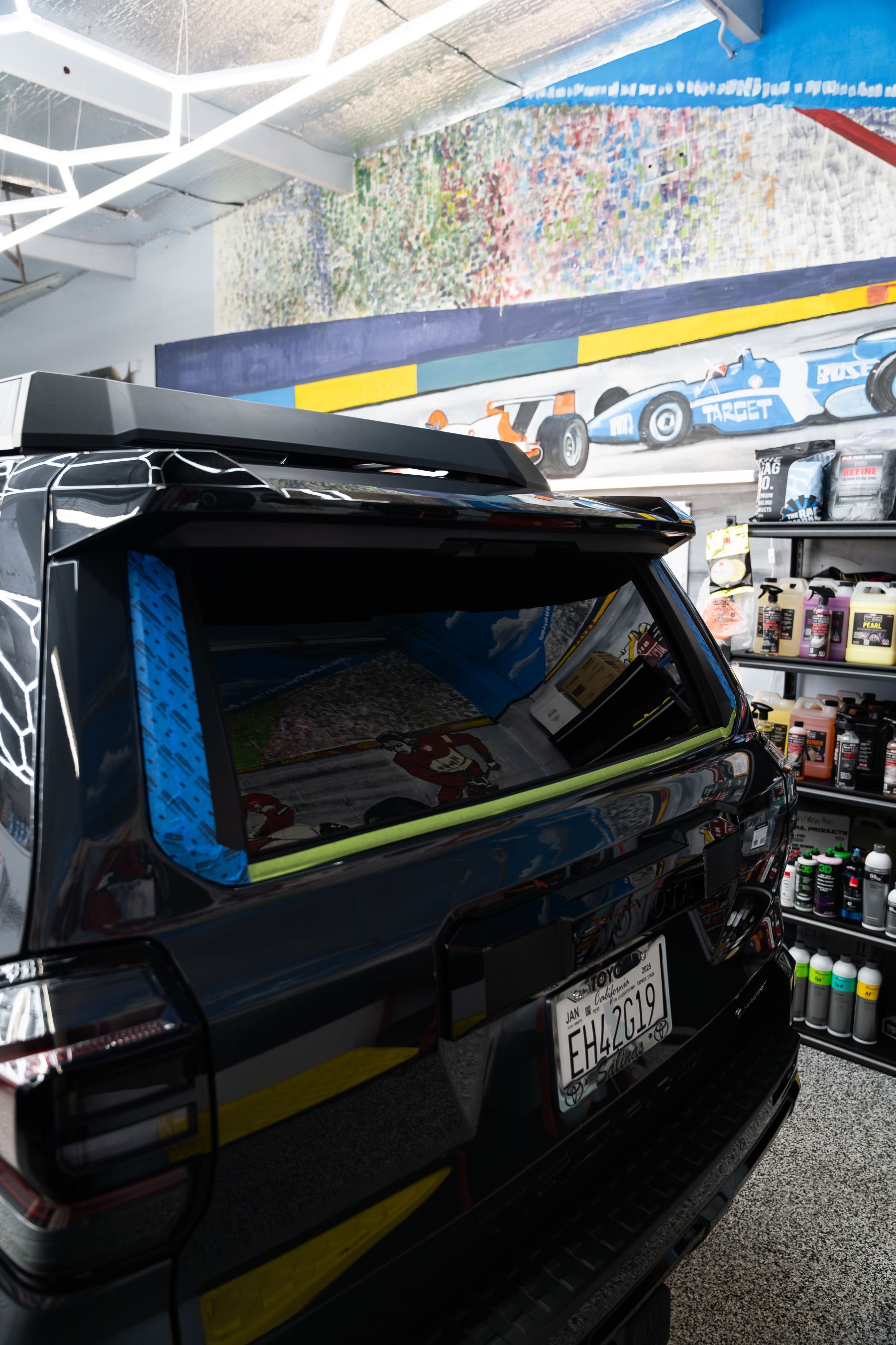 A black SUV in an auto shop has blue masking tape on its rear window frame, preparing for glass replacement or repair.