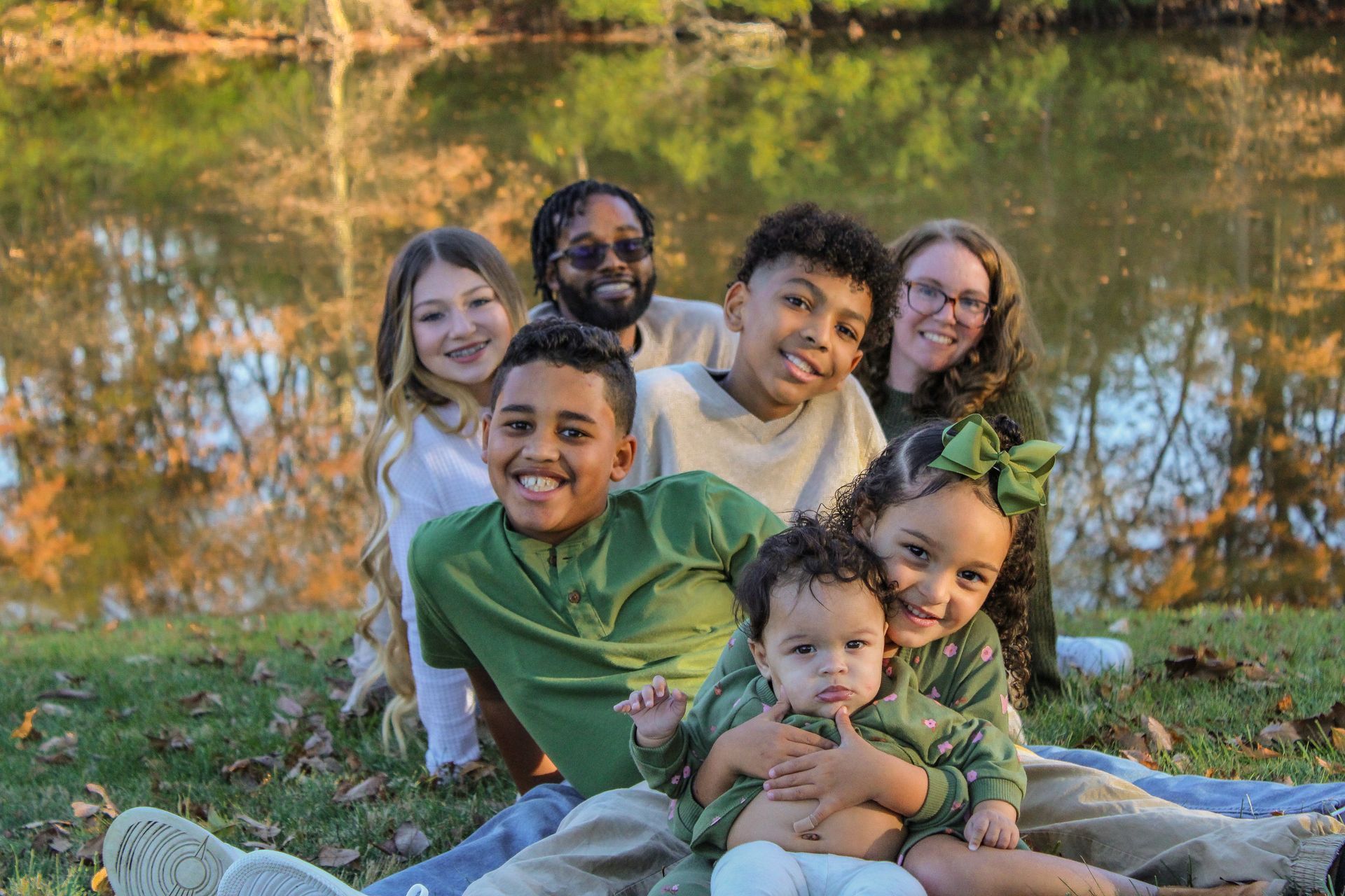 Family of seven smiles for the camera, seated by a reflective lake, wearing shades of green.