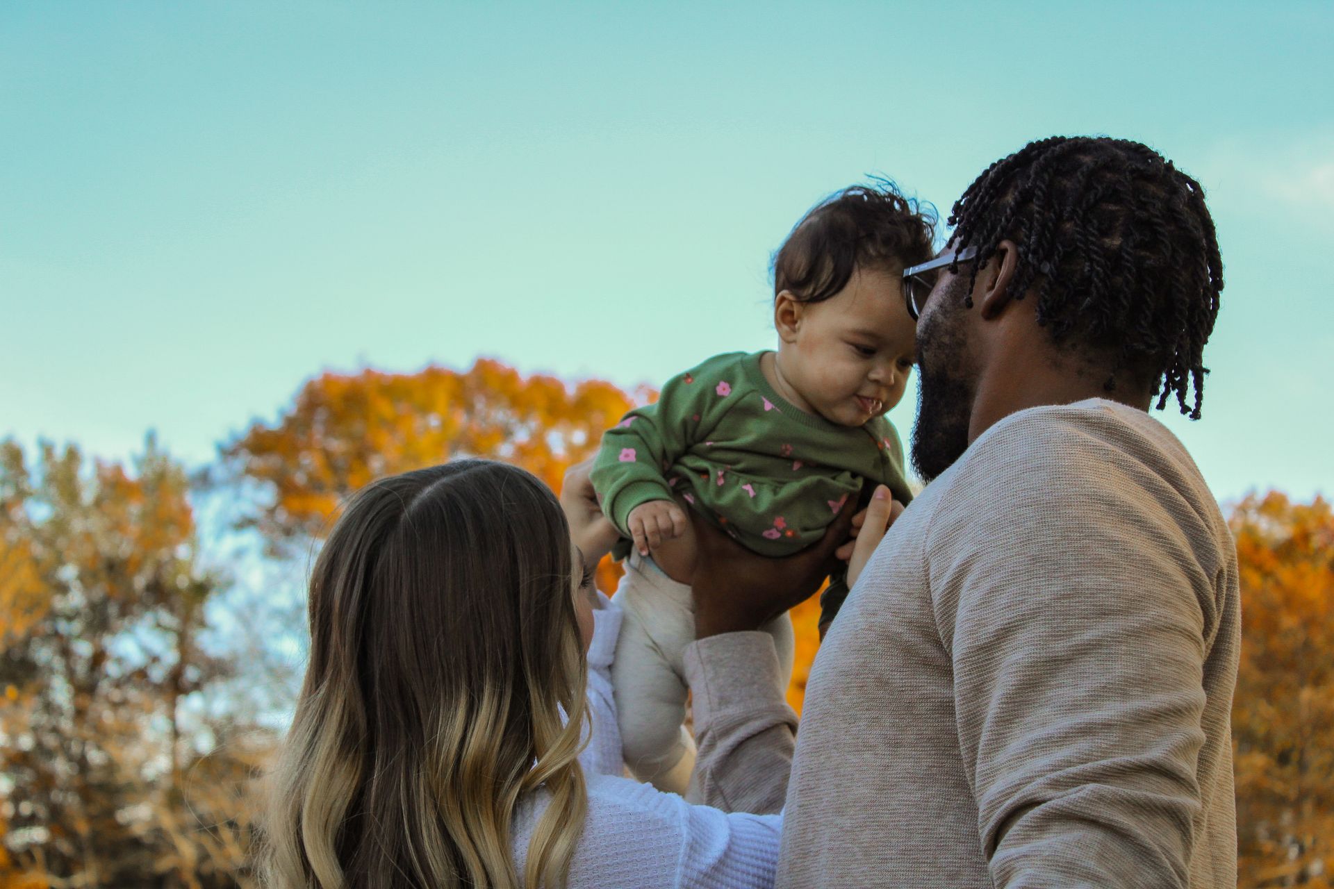 Parents lifting a baby up in the air. Autumn trees and a blue sky in the background.