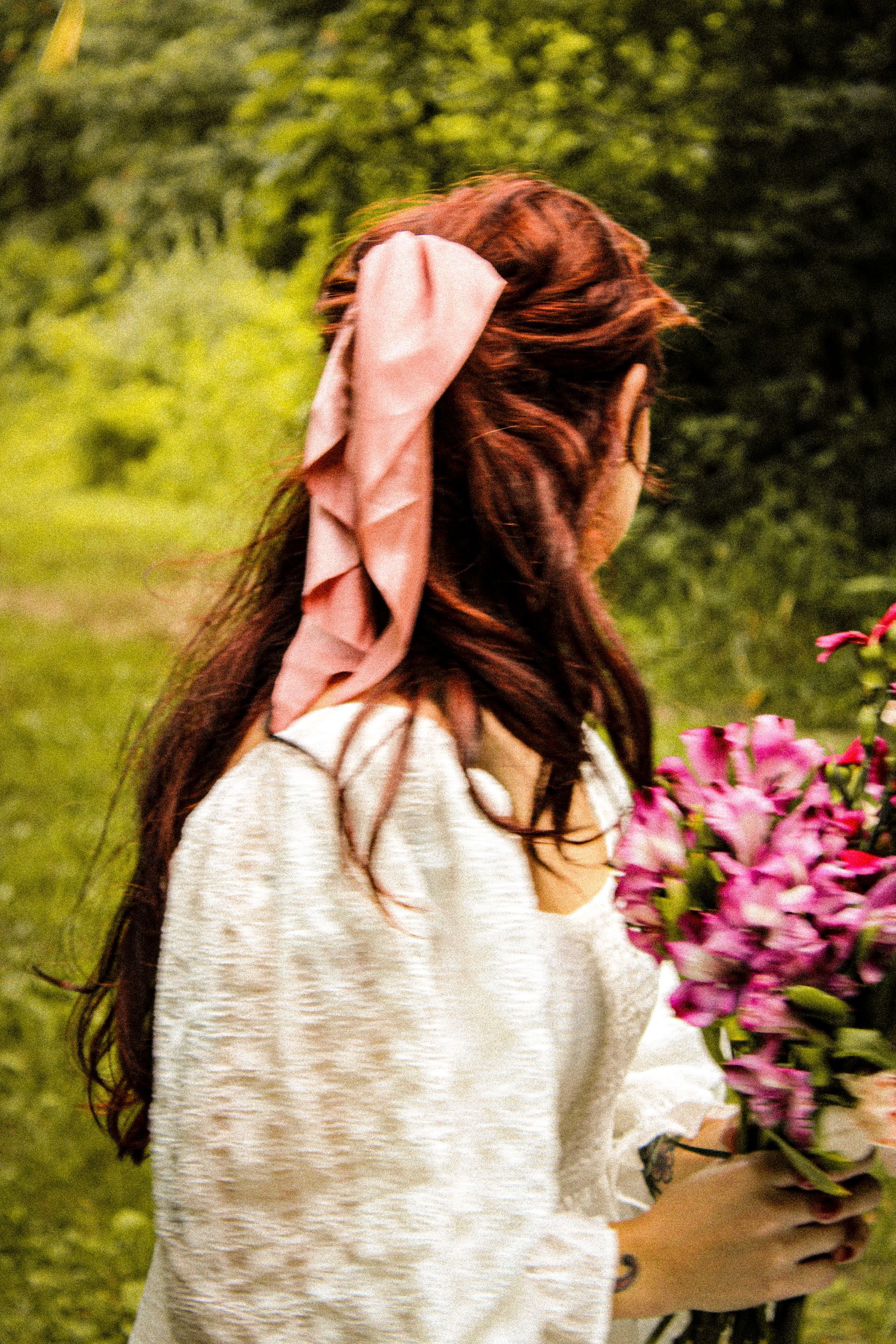 Woman with auburn hair and pink ribbon, holding pink flowers in a green field, wearing white shirt.