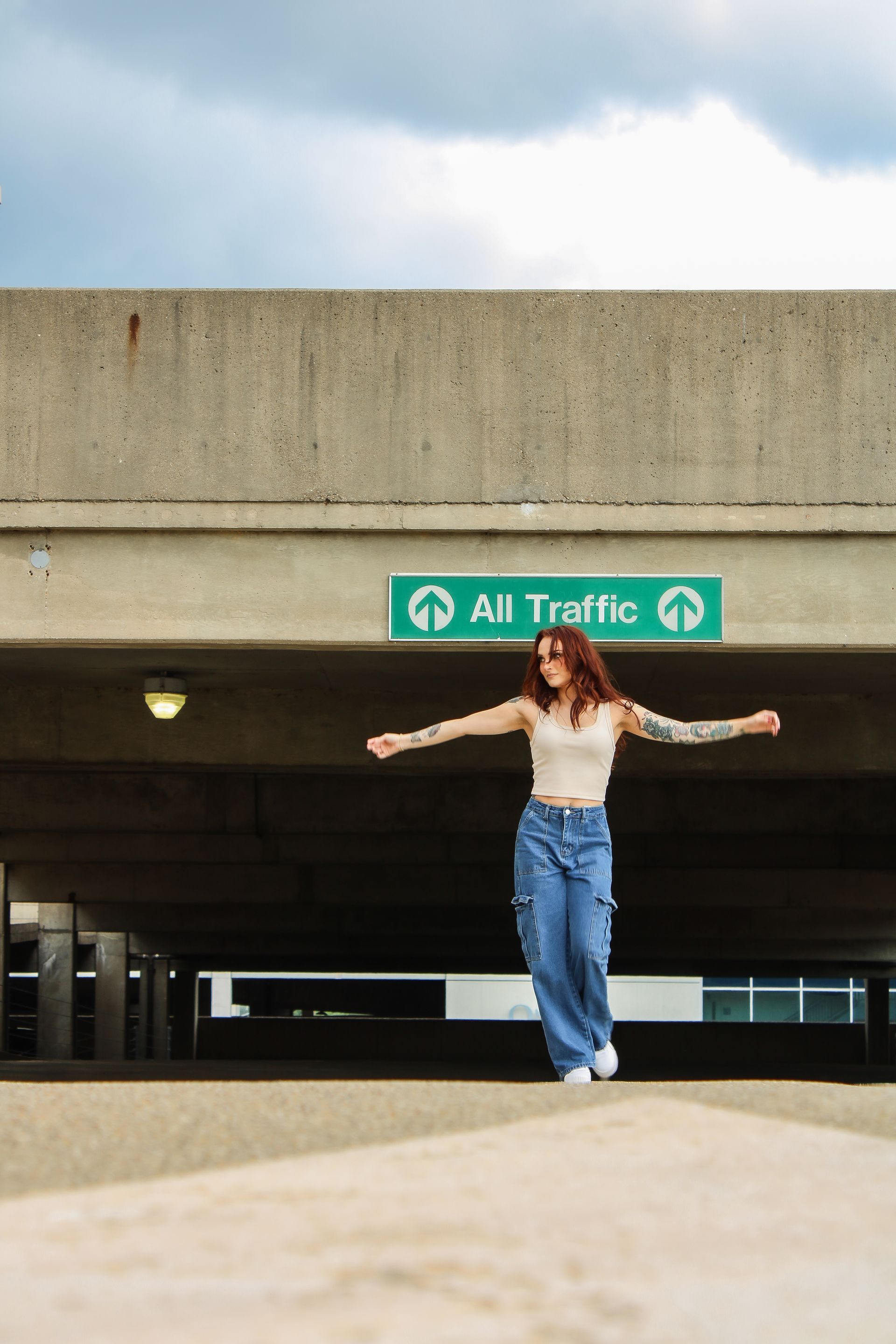 Woman with arm tattoos stands in parking garage, arms outstretched.