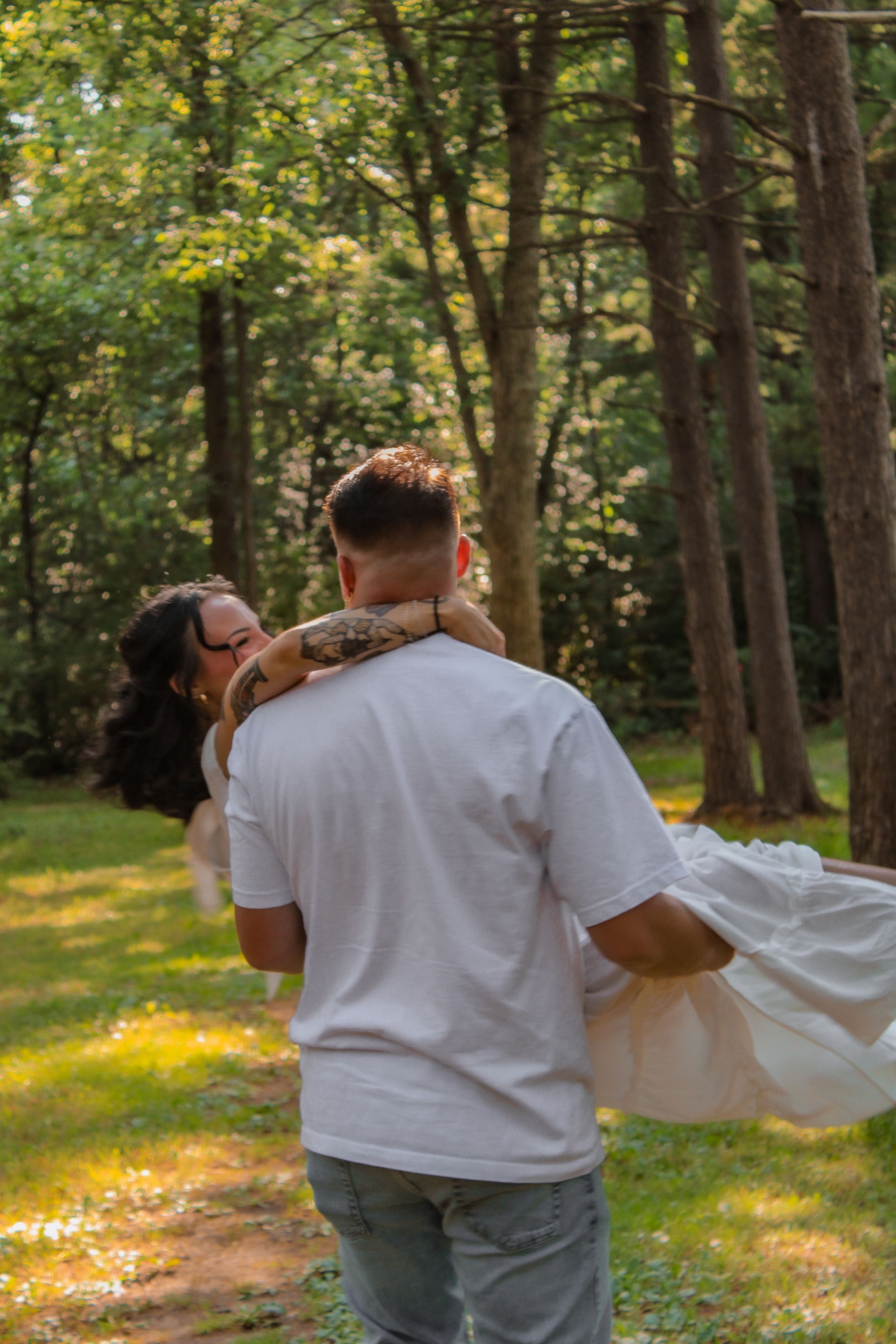 Man carrying woman in a forest, both smiling. Woman in white dress, man in white shirt and jeans.