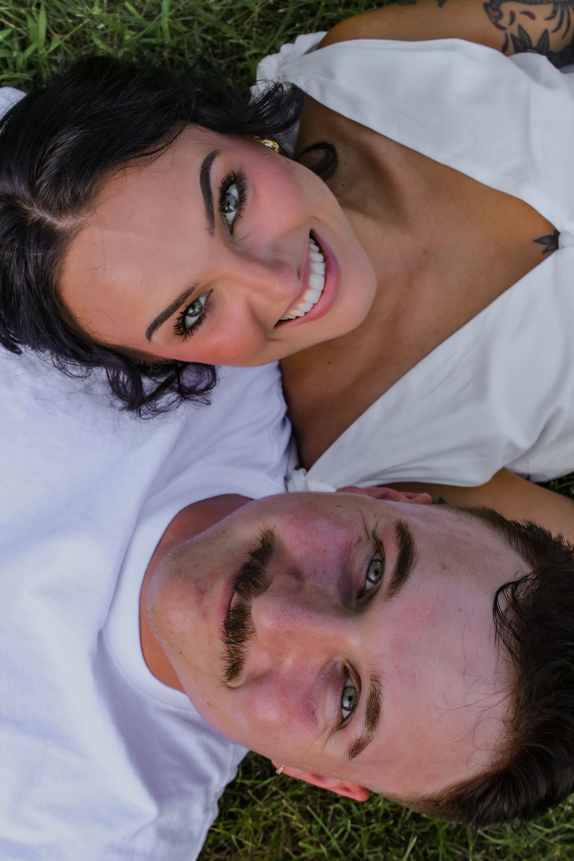 Couple smiling, lying on grass. Woman in white top, man in white shirt with mustache.