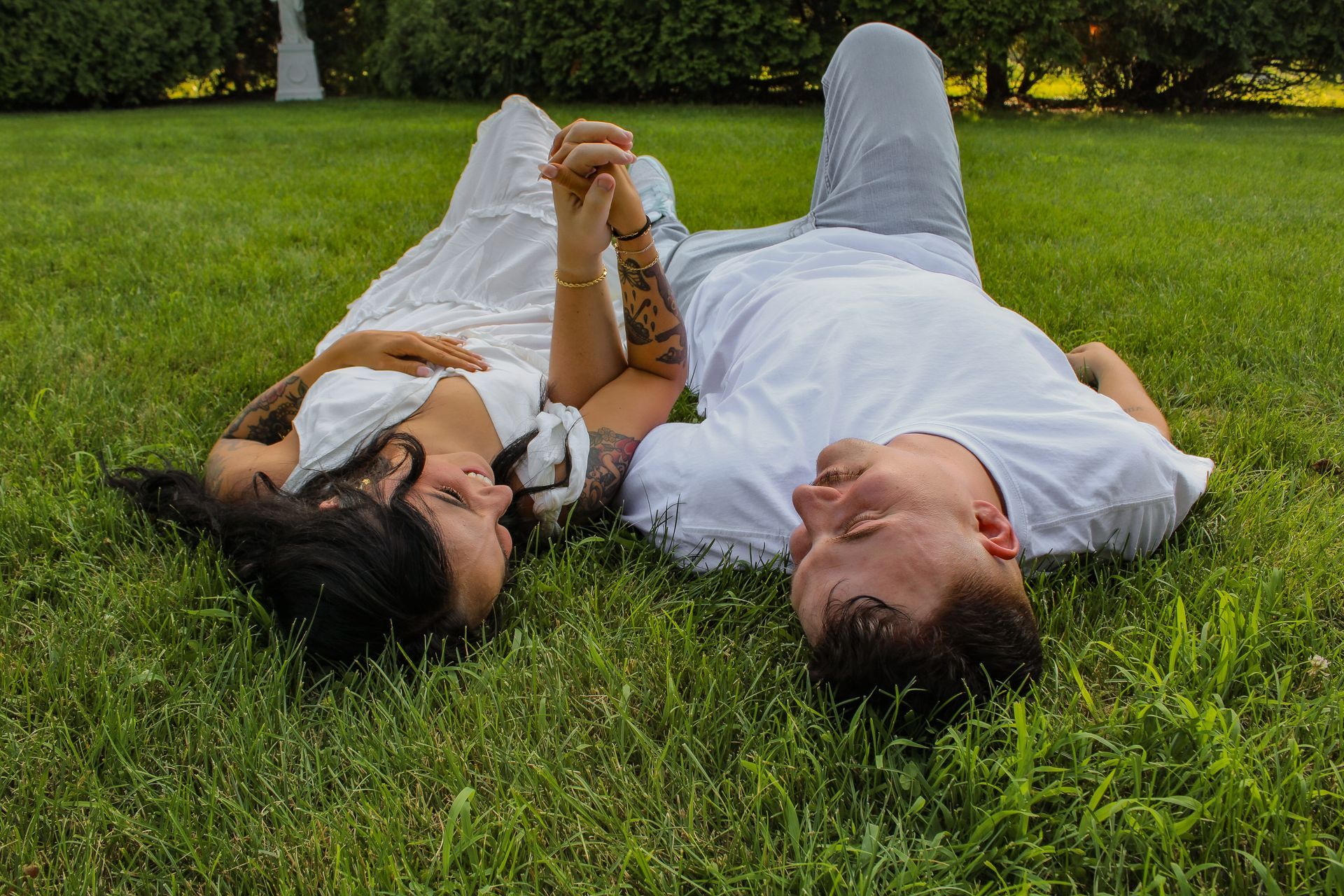 Couple lying in grass, holding hands, looking at each other. Man in white shirt and grey pants; woman in white dress.