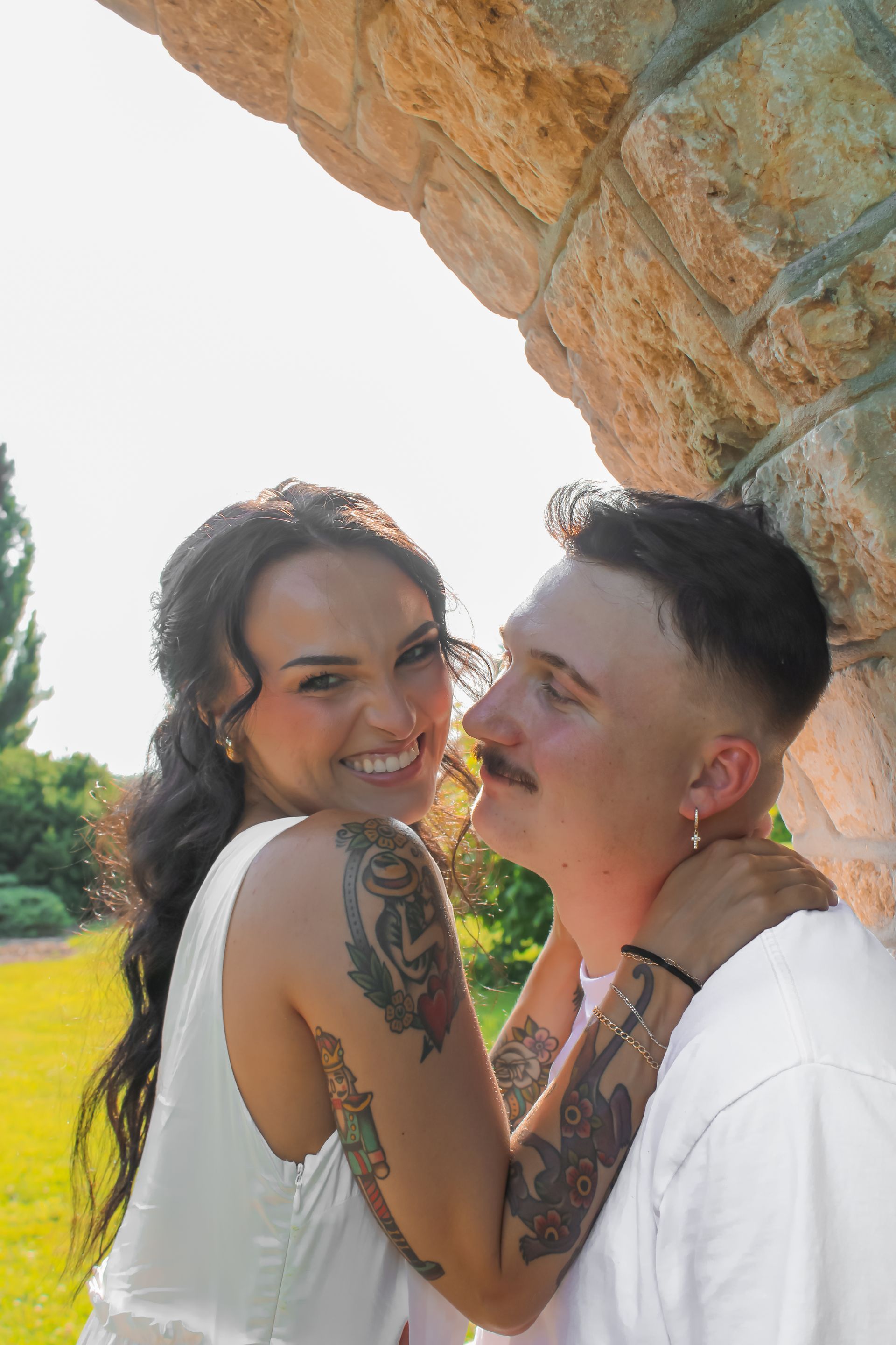 Couple smiling, hugging under stone archway. Woman with tattoos, man with mustache, sunny outdoor setting.