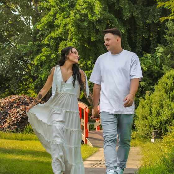 A man and a woman are walking down a path holding hands . the woman is wearing a white dress.