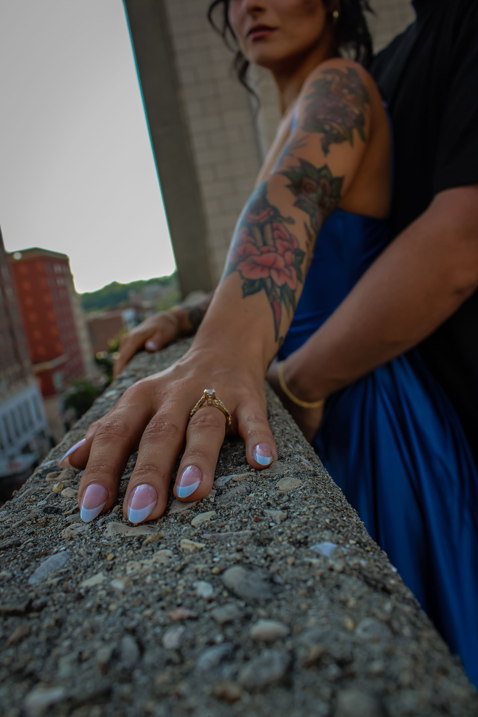 Woman with arm tattoos and blue dress, leaning on a wall; man's arm around her.