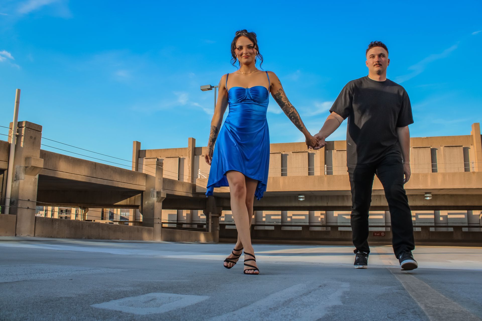 Couple holding hands on a rooftop parking lot; woman in blue dress, man in black t-shirt.