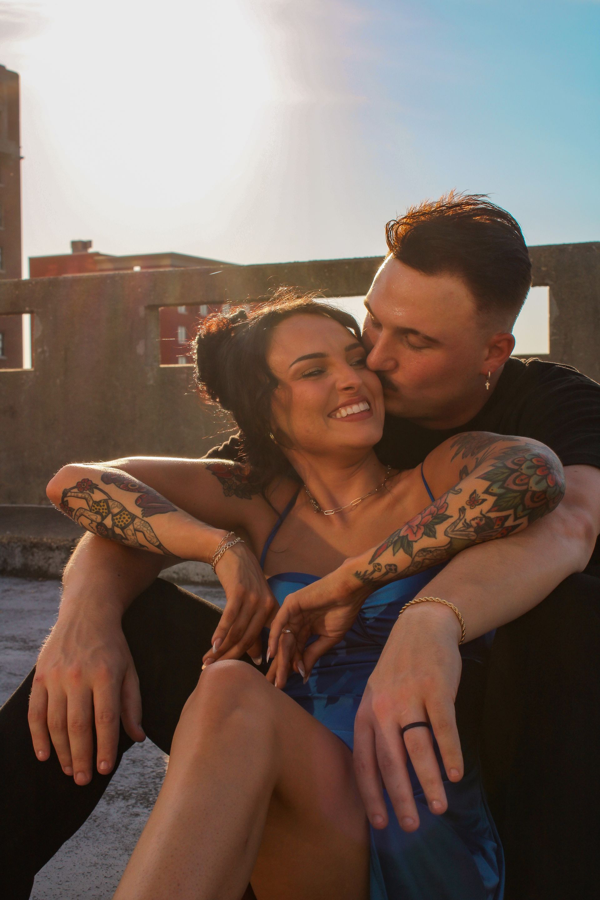 Couple embraces on rooftop; man kisses woman's cheek. Golden sunlight, tattoos, blue dress.