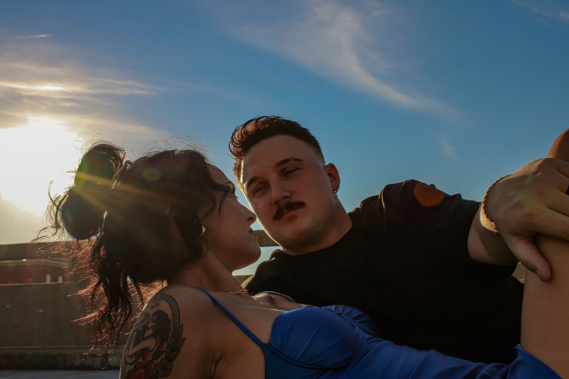 Man and woman embracing outdoors, sky background. He has a mustache, she wears a blue dress.
