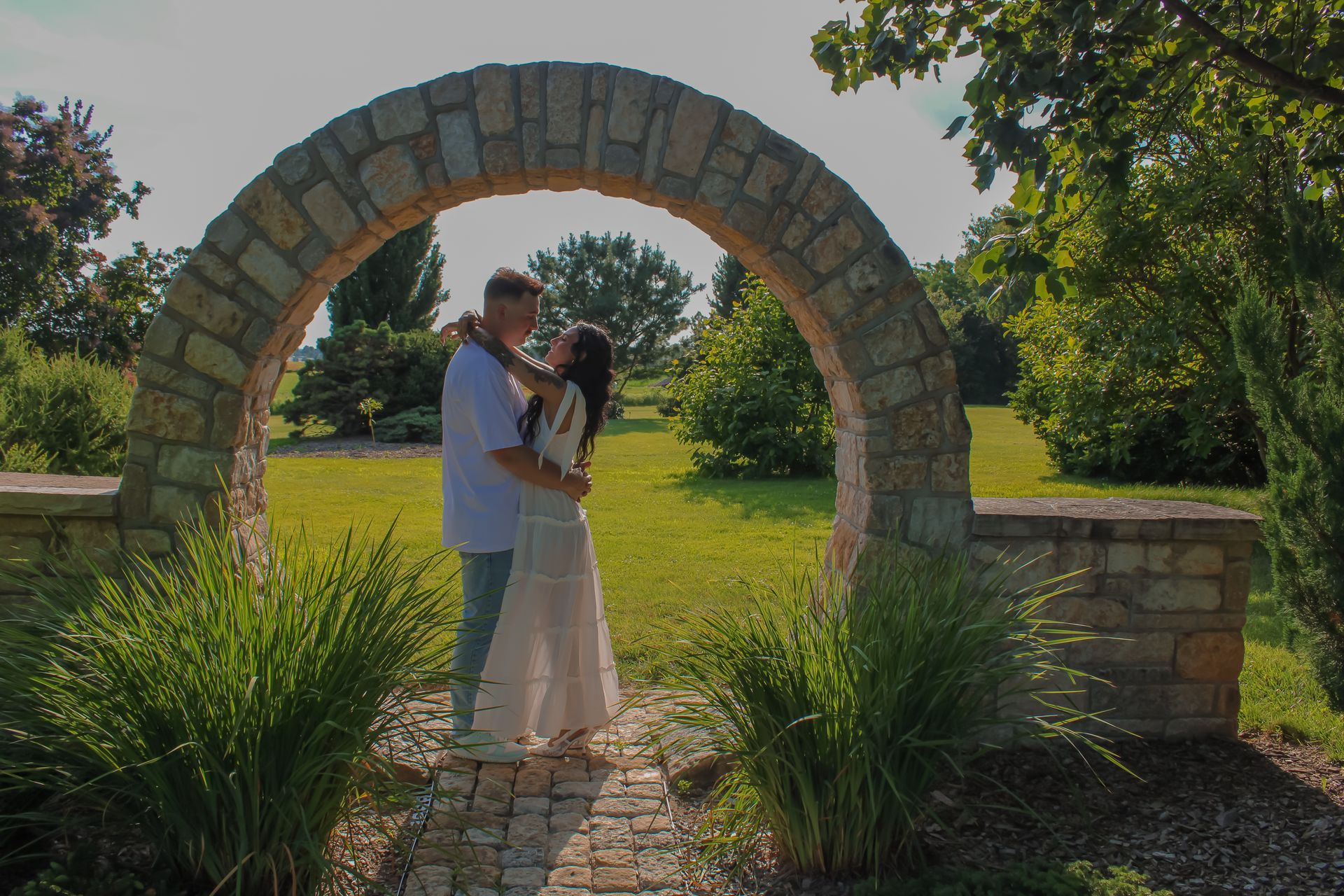 Couple embracing under a stone archway in a sunny garden.