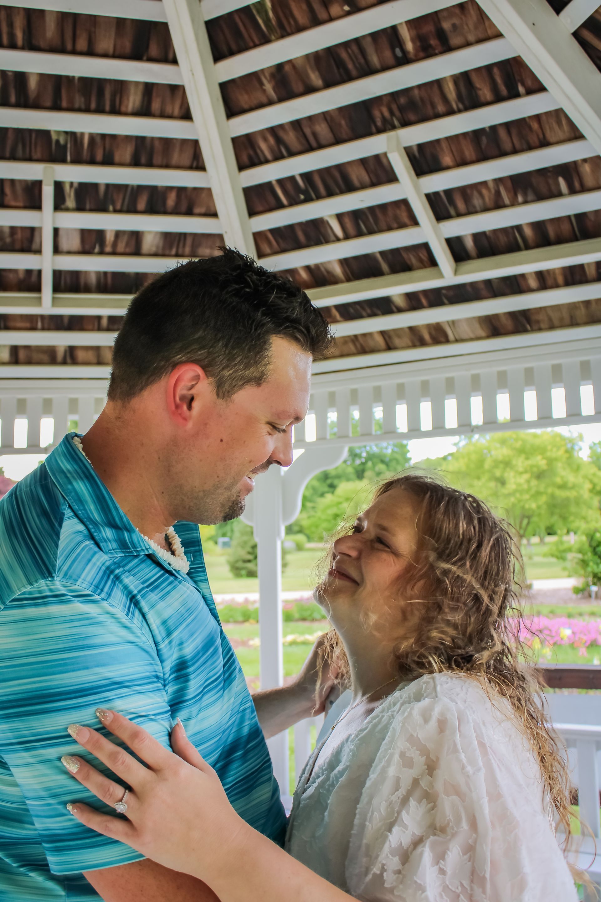 Couple smiling and embracing in a white gazebo, looking at each other. Blue shirt, white dress, sunny outdoor background.